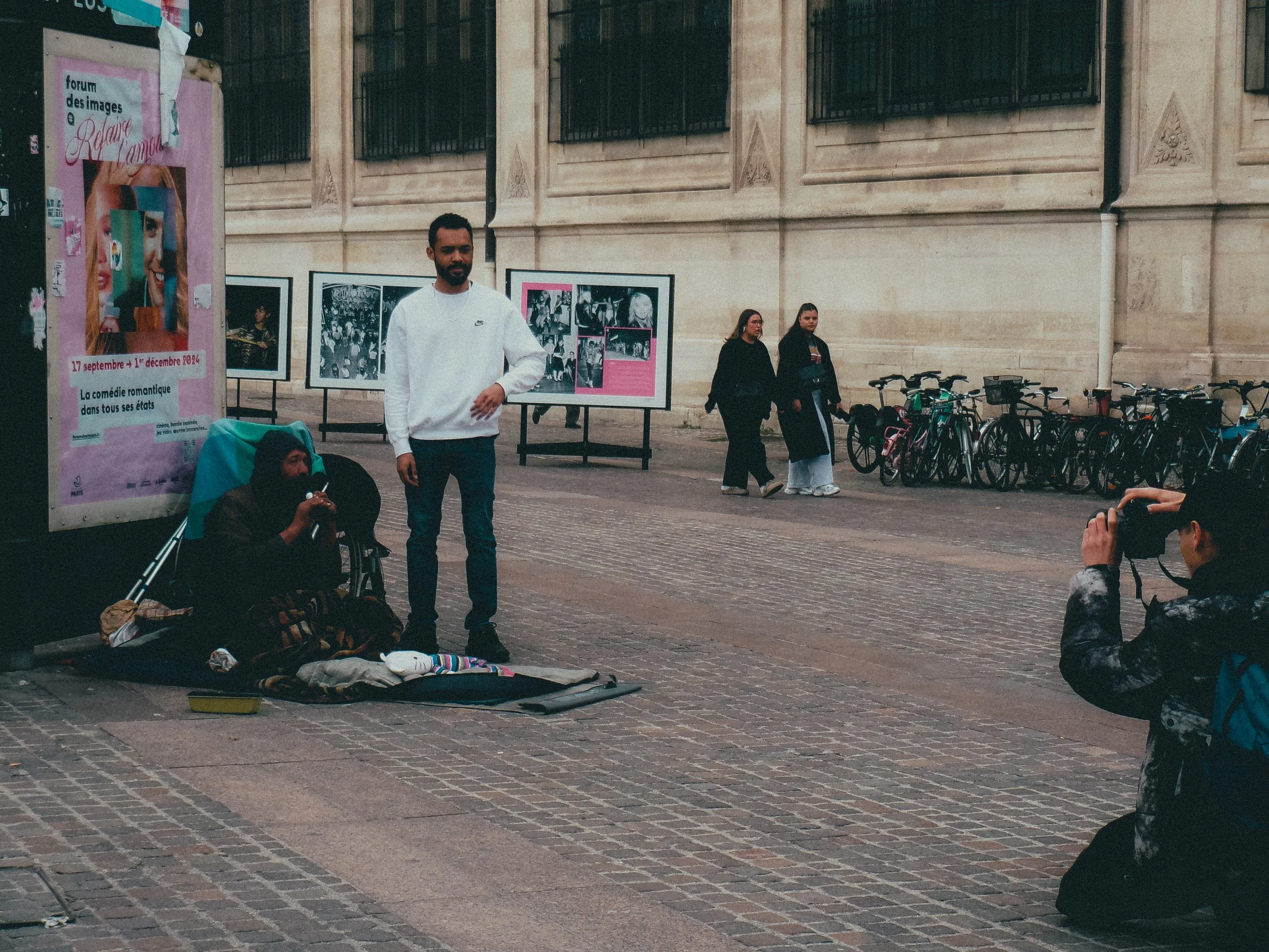 Un homme assis par terre avec un microphone, à côté d'une femme dormant avec une canne, à côté d'un panneau d'affichage. Deux femmes marchent en arrière-plan et un jeune homme prend une photo.