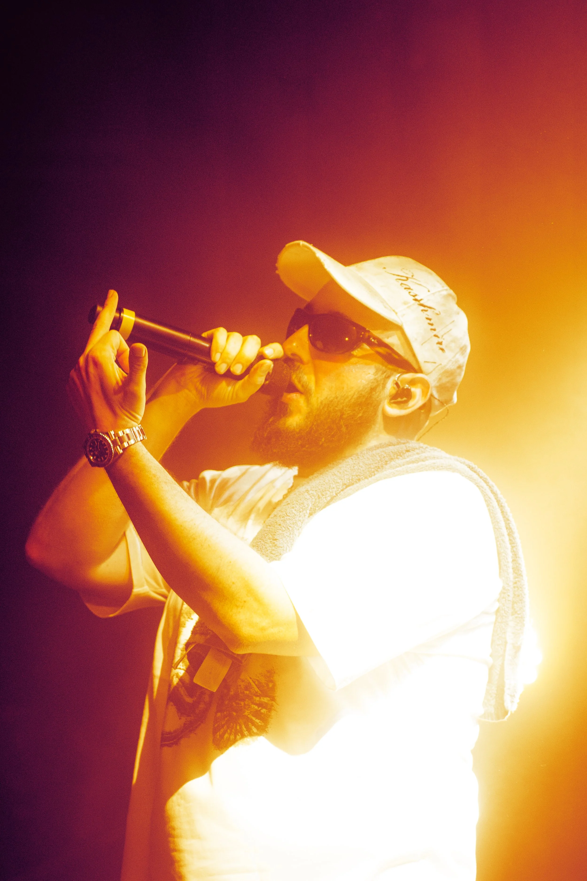 Un homme portant une casquette, des lunettes de soleil et une montre, chantant ou parlant dans un microphone, avec un éclairage chaud orange.