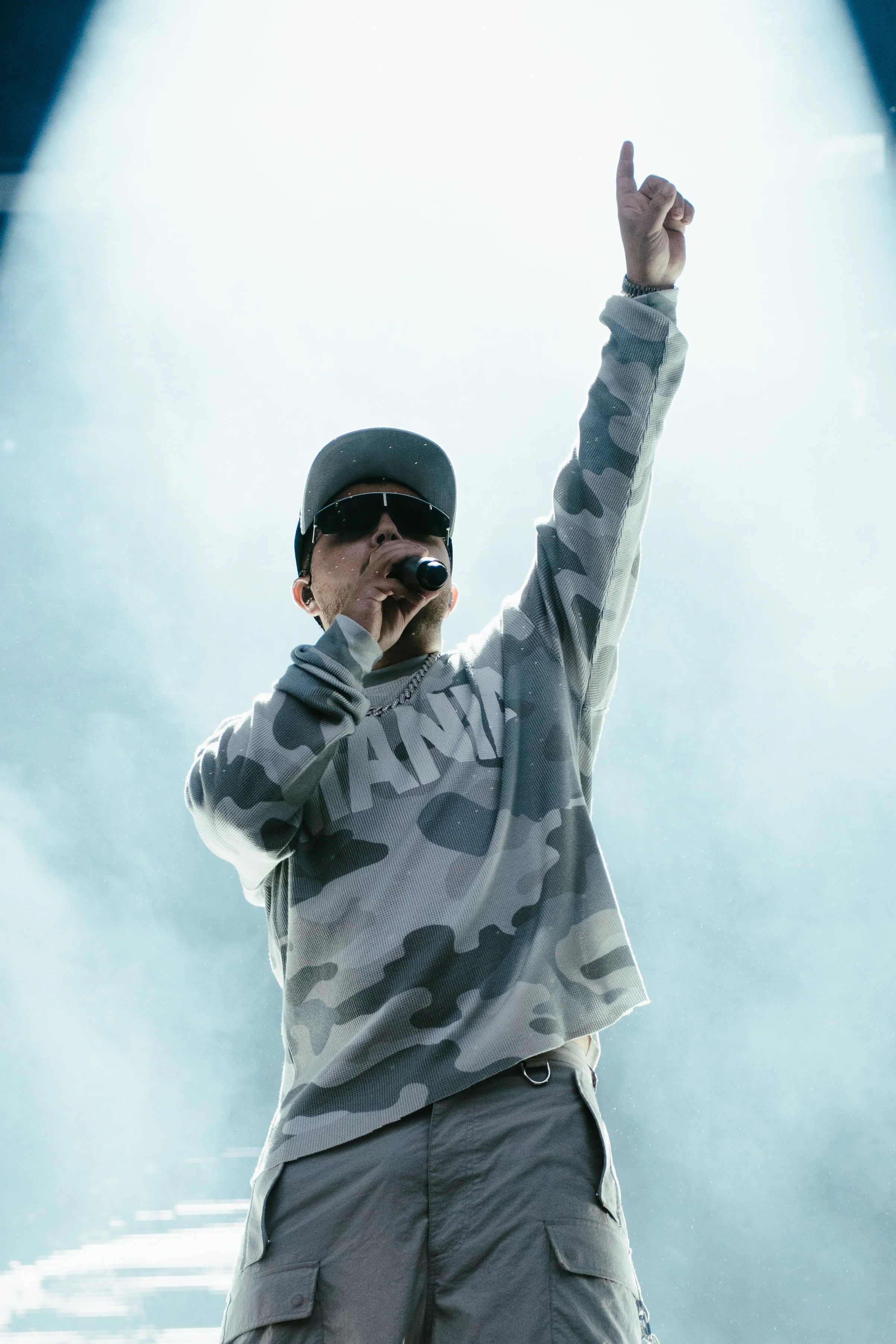 Un homme portant des lunettes de soleil, un chapeau et un sweat-shirt camouflage chante en tenant un microphone, avec un bras levé, sur scène sous des lumières vives.
