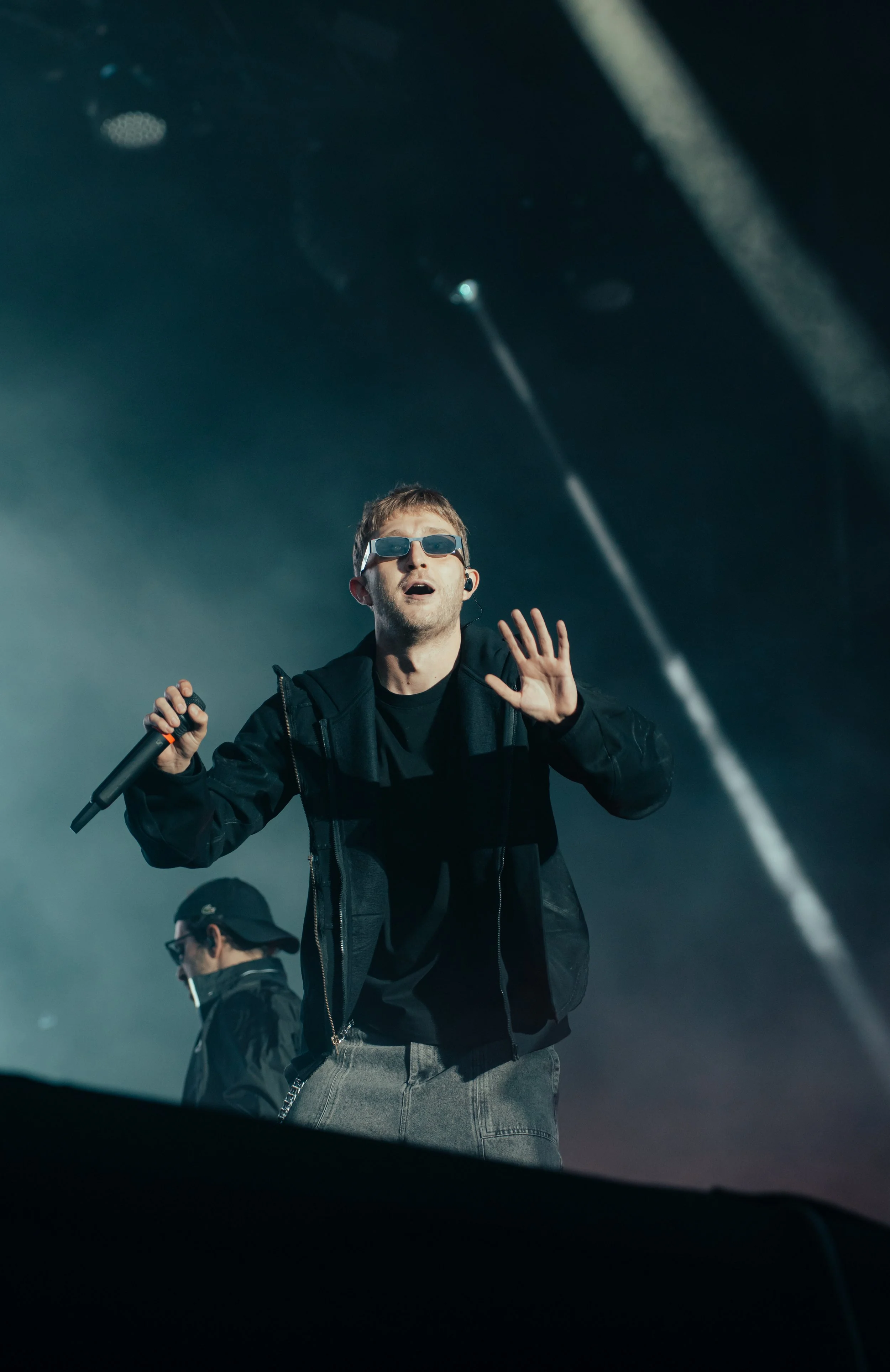 Un homme portant des lunettes de soleil, une veste noire, et tenant un microphone, chante ou parle sur scène, avec un autre musicien en arrière-plan.
