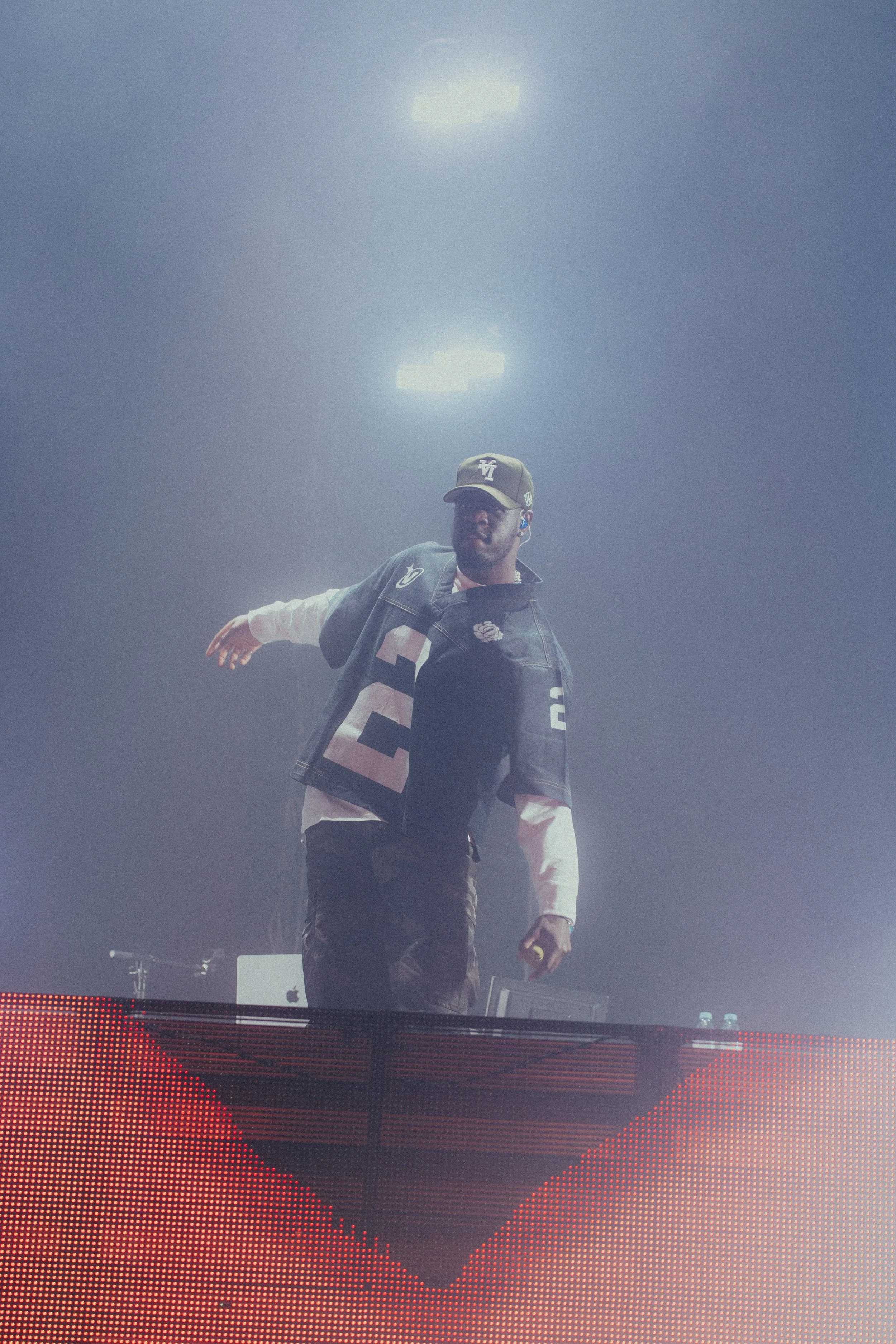 Un DJ portant une casquette et un sweat-shirt joue lors d'un concert, entouré de lumières et de fumée, derrière une table avec du matériel de musique.