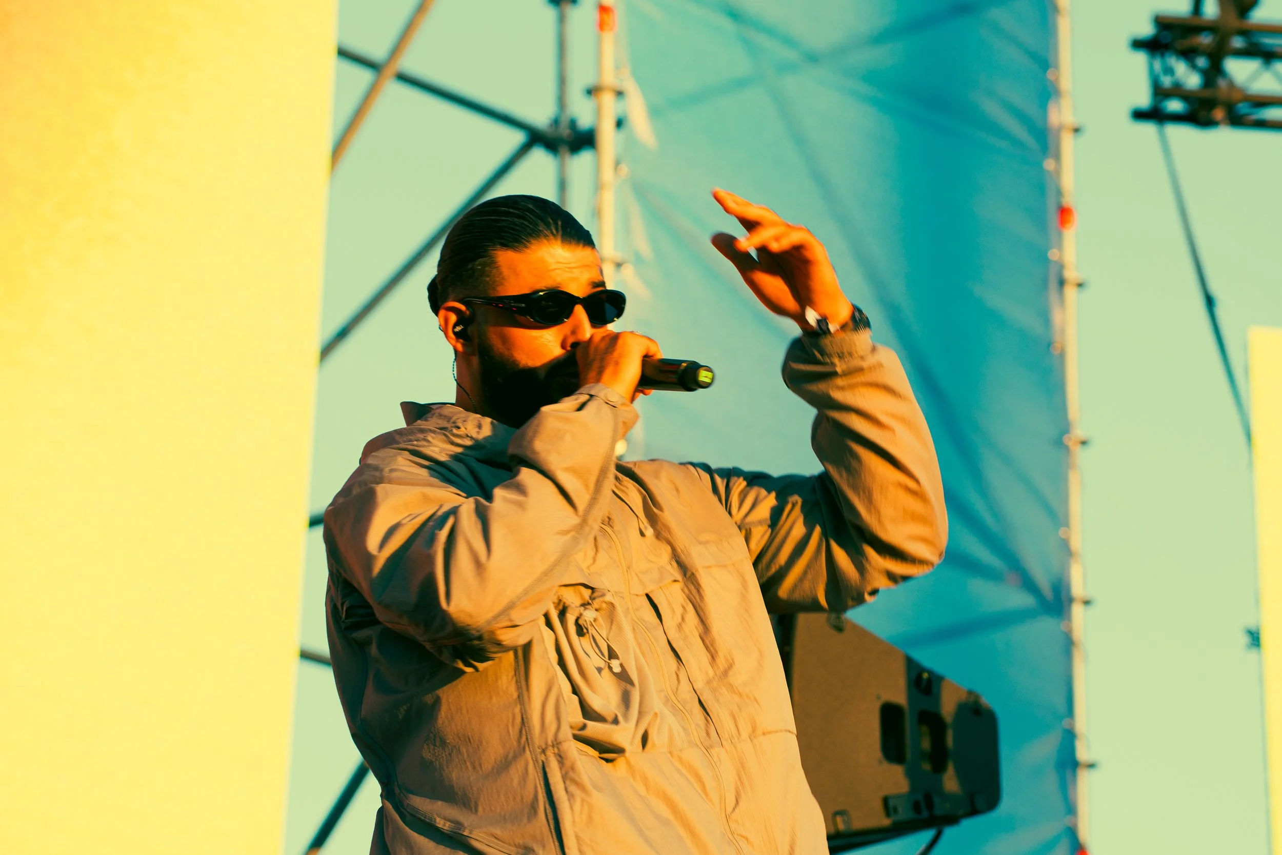 Un homme avec des lunettes de soleil, une barbe et un foulard noir, portant une veste beige, tient un micro en main tout en faisant un geste avec l'autre main sur une scène extérieure.