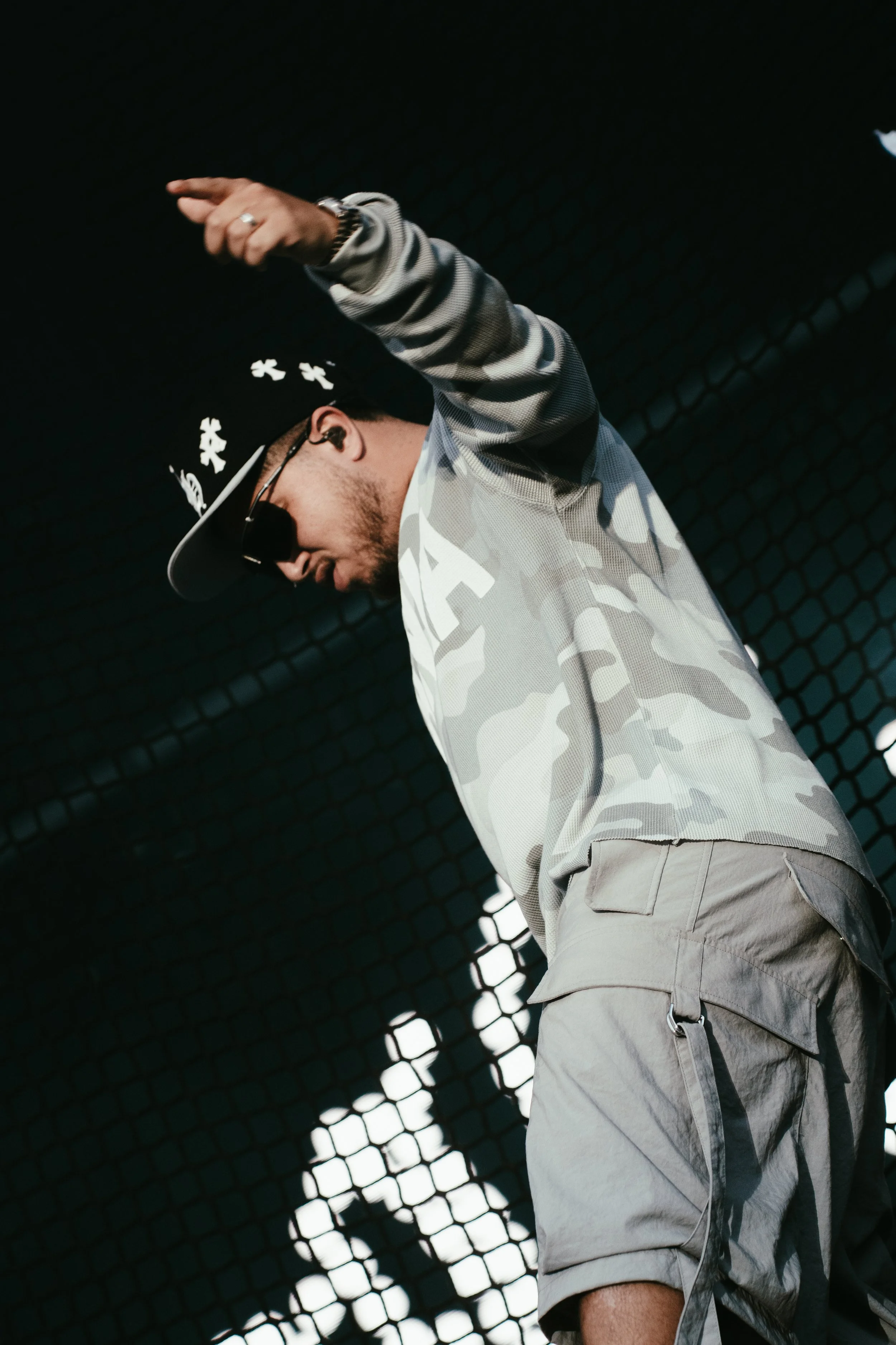Un homme portant un couvre-chef noir, des lunettes de soleil, une veste à manches longues à motifs camouflage gris et beige, et un short beige, semble danser ou performer sur scène contre un fond noir avec un motif en grille.