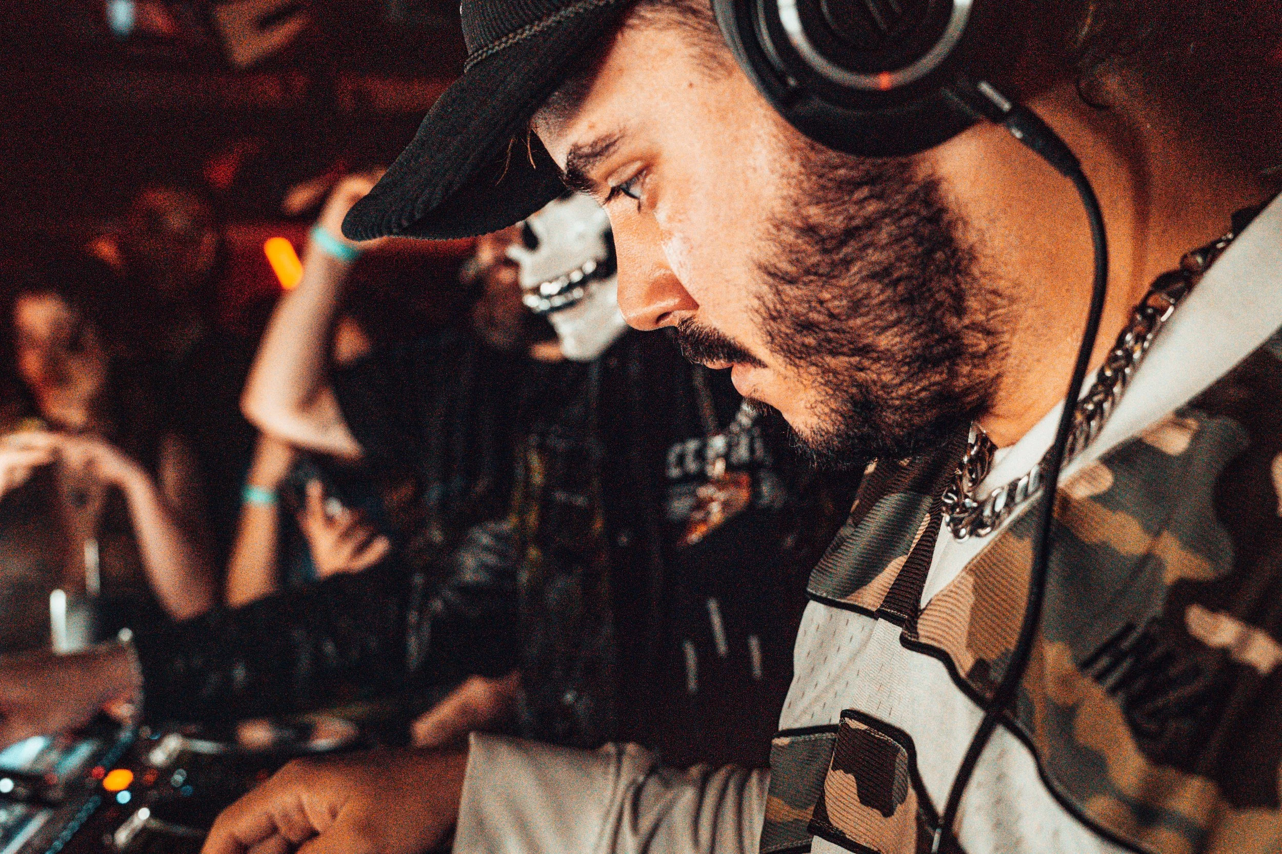 Un DJ avec une barbe qui porte un casque audio, un chapeau noir et un collier, en train de mixer lors d'une fête ou d'un événement, avec un public en arrière-plan.