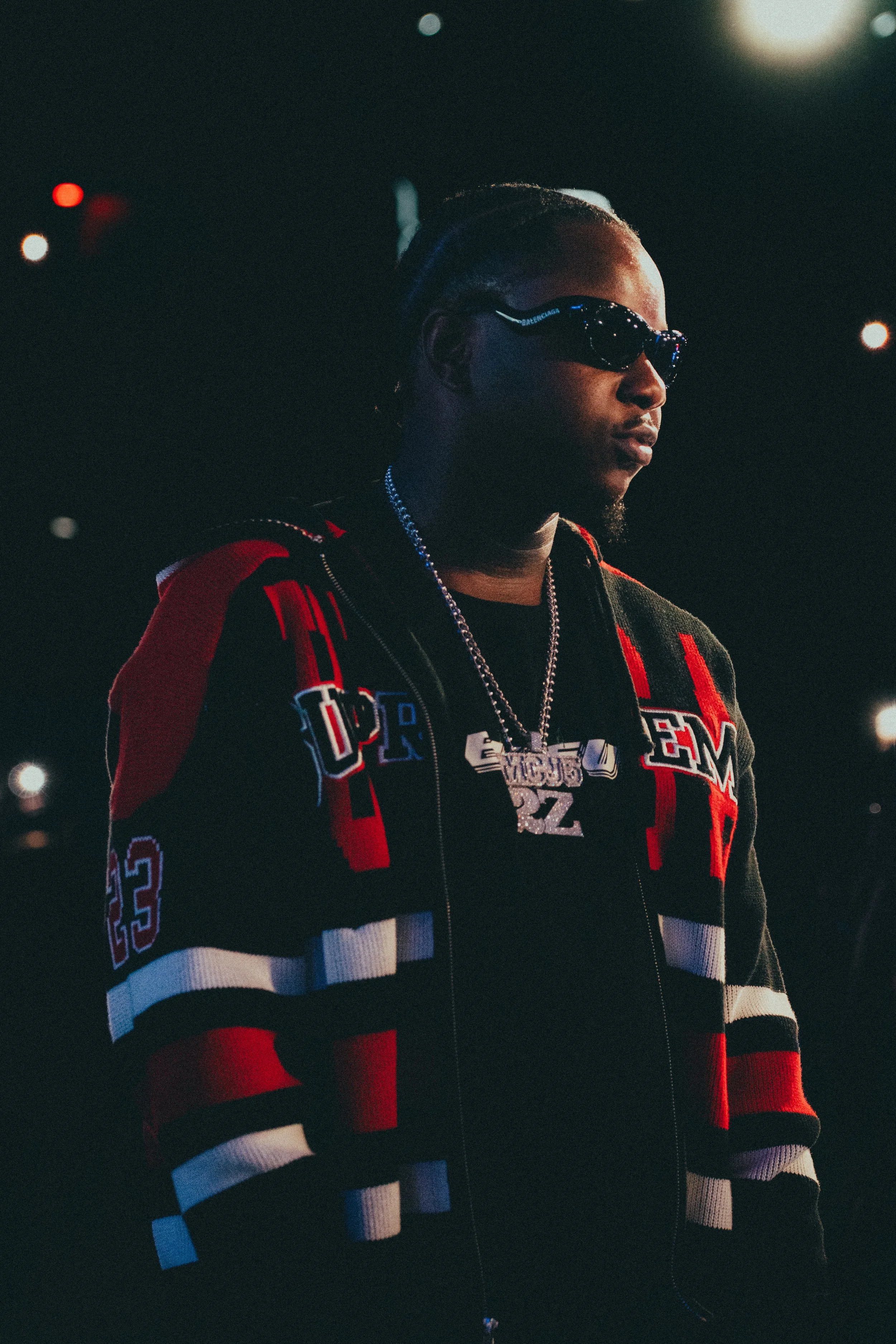 Un homme portant des lunettes de soleil, un sweat à capuche noir et un veste colorée noire, rouge, blanc et gris, dans un environnement sombre avec des lumières floues en arrière-plan.
