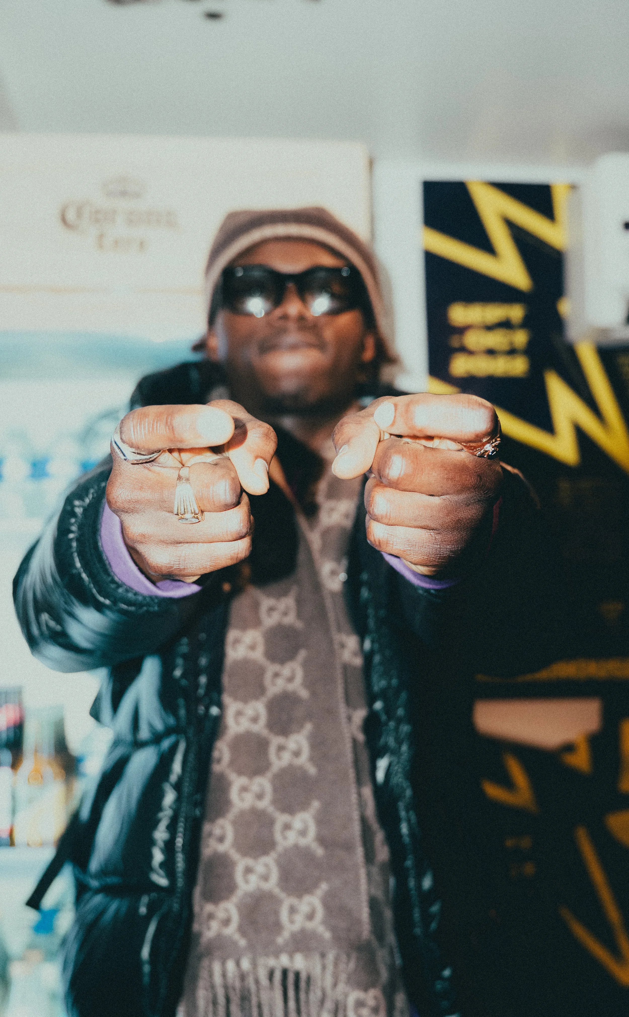 Une personne portant des lunettes de soleil, un bonnet, une veste noire, et une écharpe, pointant directement la caméra.