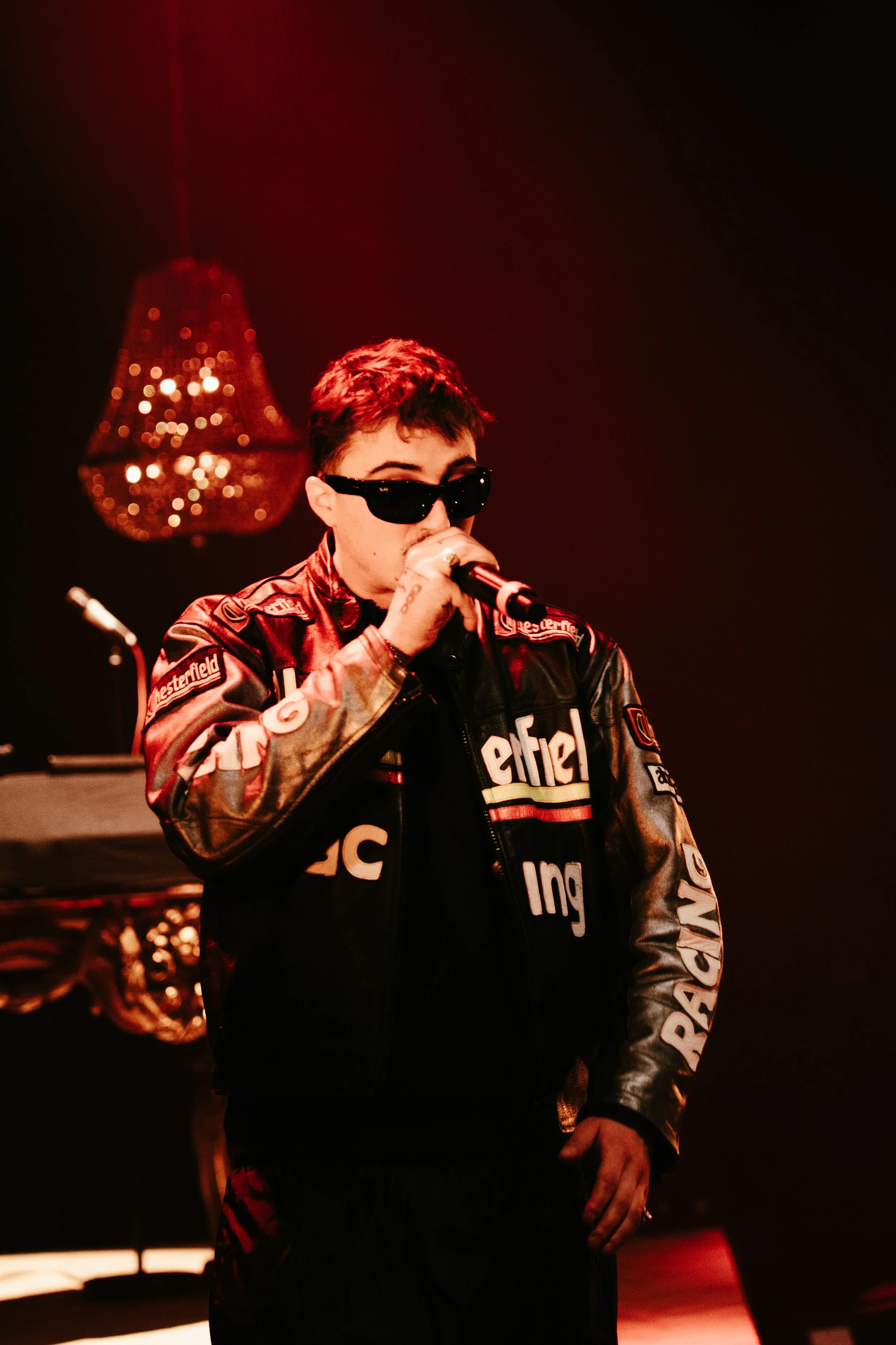 Une personne portant des lunettes noires et une veste en cuir avec des logos, tenant un micro et faisant une performance sur scène avec un éclairage rouge, arrière-plan flou avec un lustre.