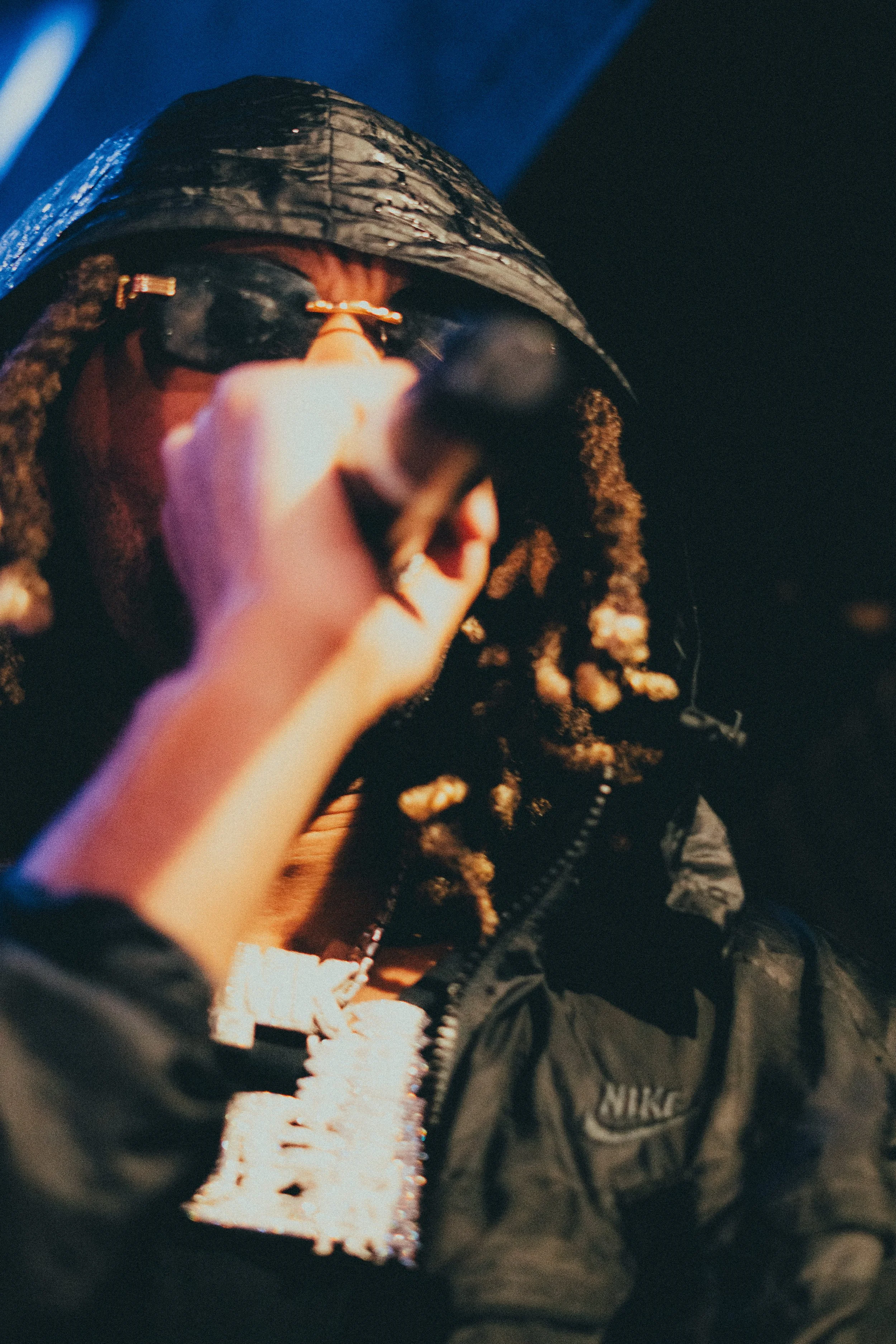 Personne portant un sweat Nike noir avec un manteau noir et un chapeau noir, fumant une cigarette dans une pièce sombre.