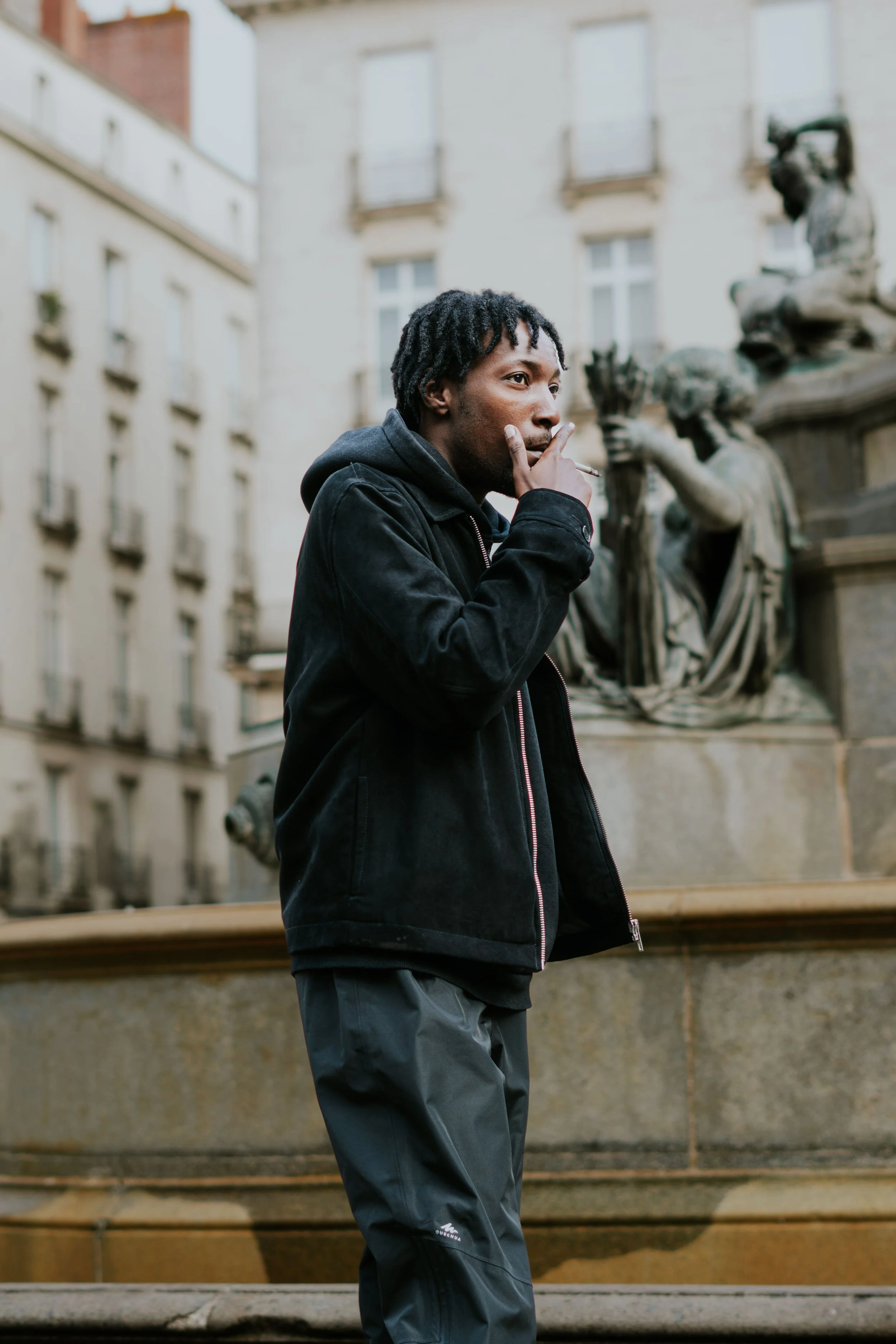 Jeune homme en vêtements décontractés, tenant un cigarette à la main près d'une fontaine et d'une sculpture dans une vieille ville européenne.