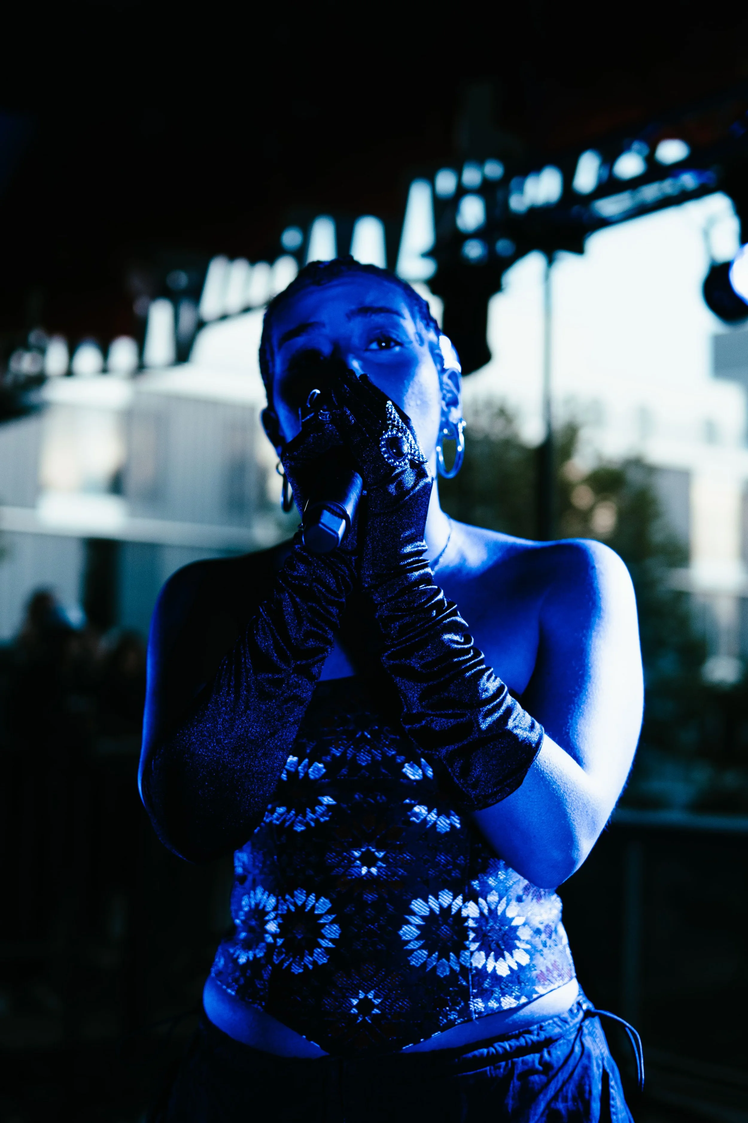 Une jeune femme chante dans un casque au microphone, portant des gants longs en satin noir, sous un éclairage bleu, avec un arrière-plan flou de bâtiments résidentiels.