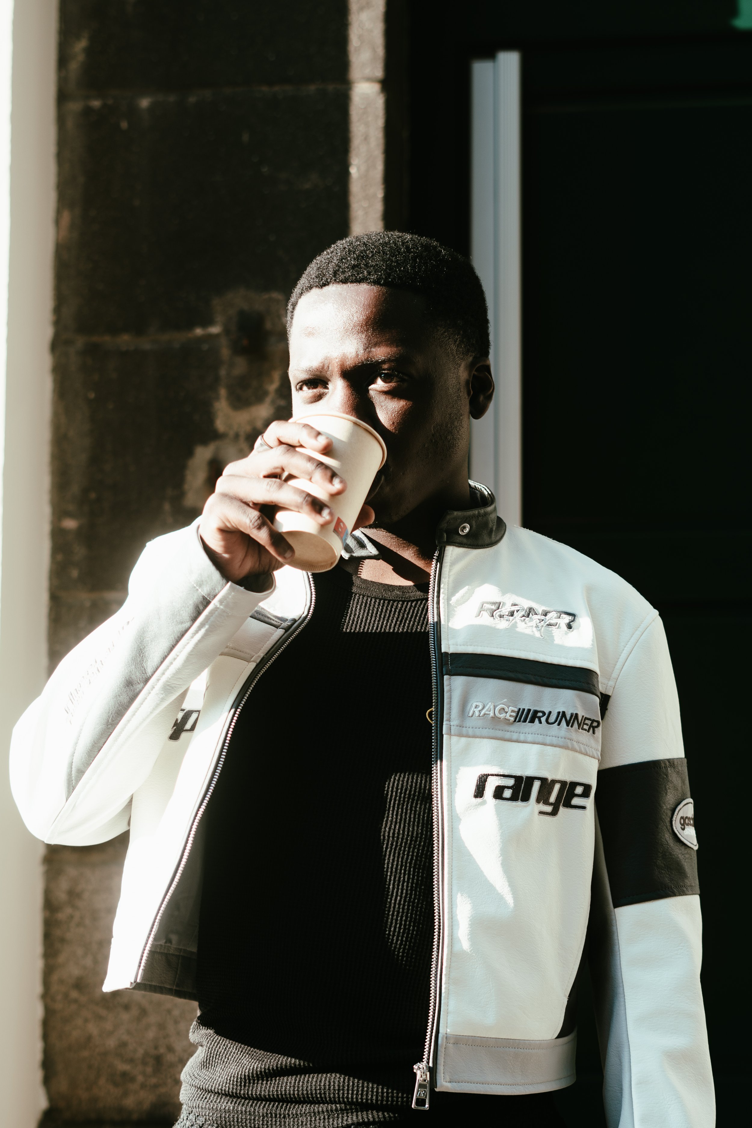 Un homme noir portant une veste de moto blanche avec des patches, buvant dans un gobelet en carton, à la lumière du soleil.