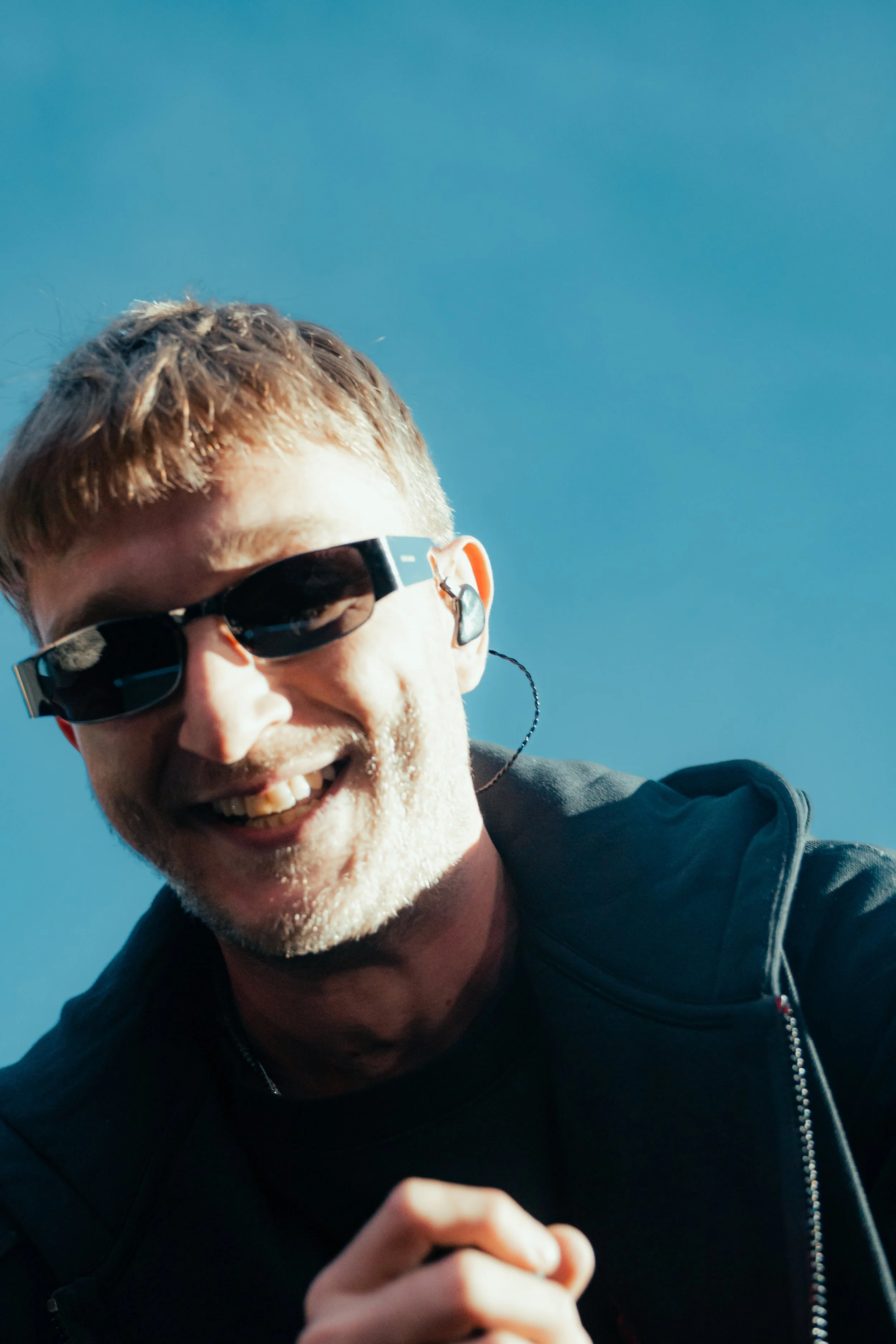 Un homme souriant portant des lunettes de soleil et un écouteur sans fil, avec un ciel bleu en arrière-plan.