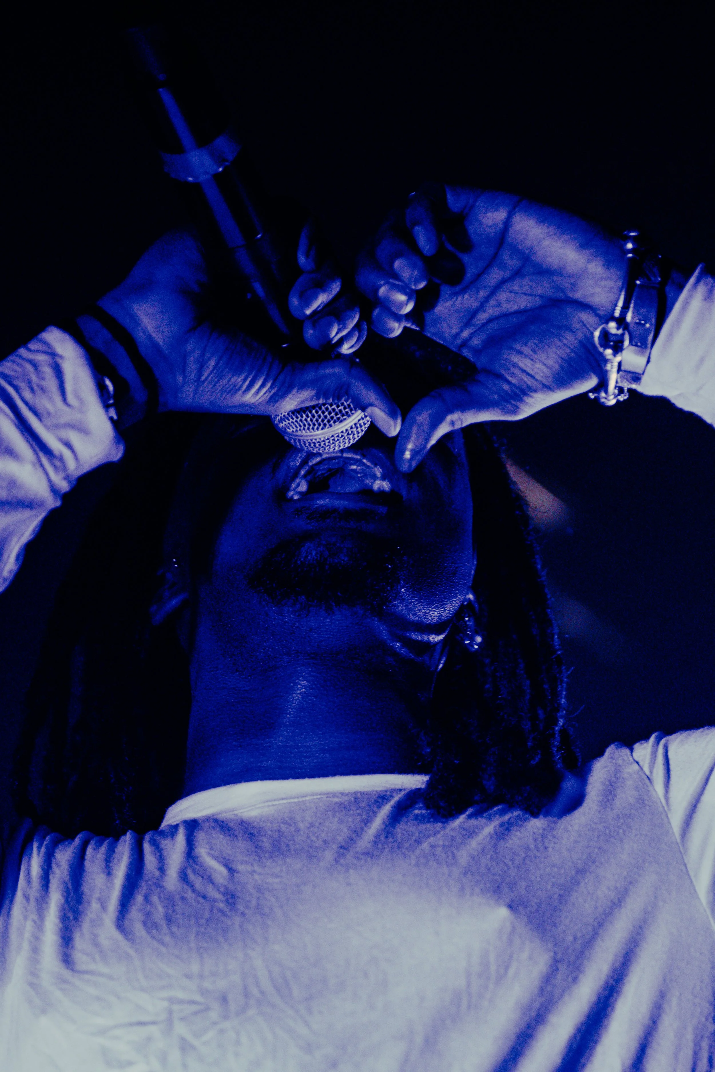 Un chanteur avec dreadlocks, tenant un microphone, chante en regardant vers le haut dans une lumière bleue.