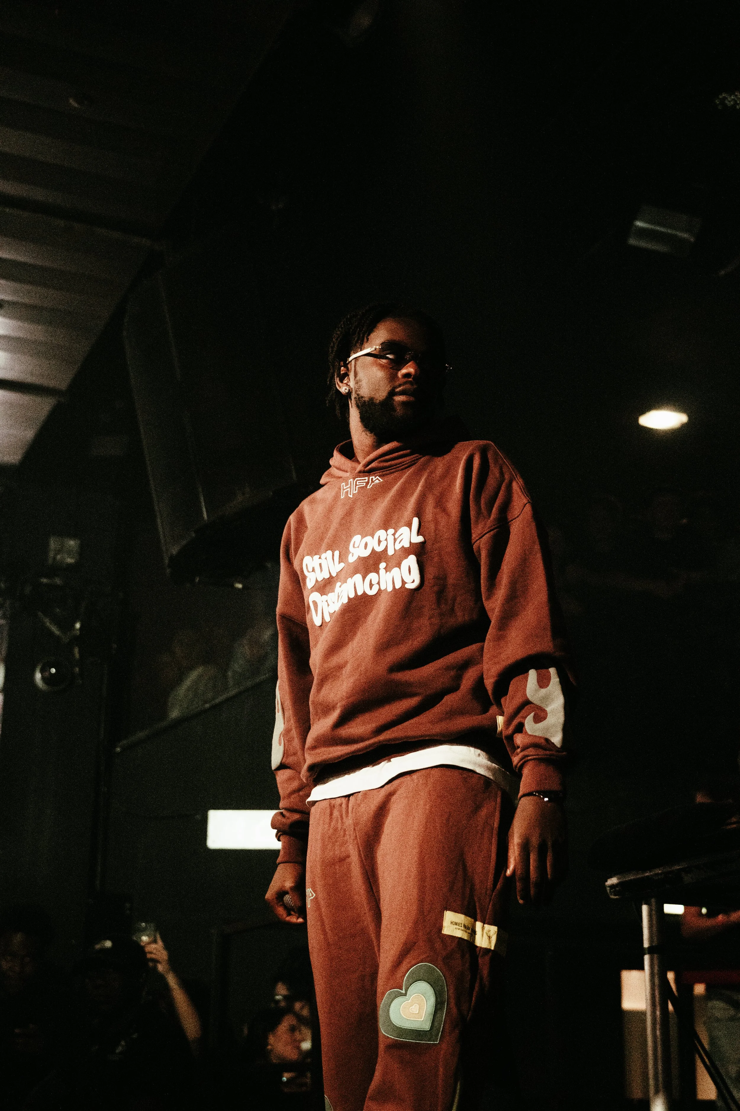 Un homme portant une veste à capuche marron avec des inscriptions blanches, debout dans une ambiance sombre, probablement à un événement ou spectacle.