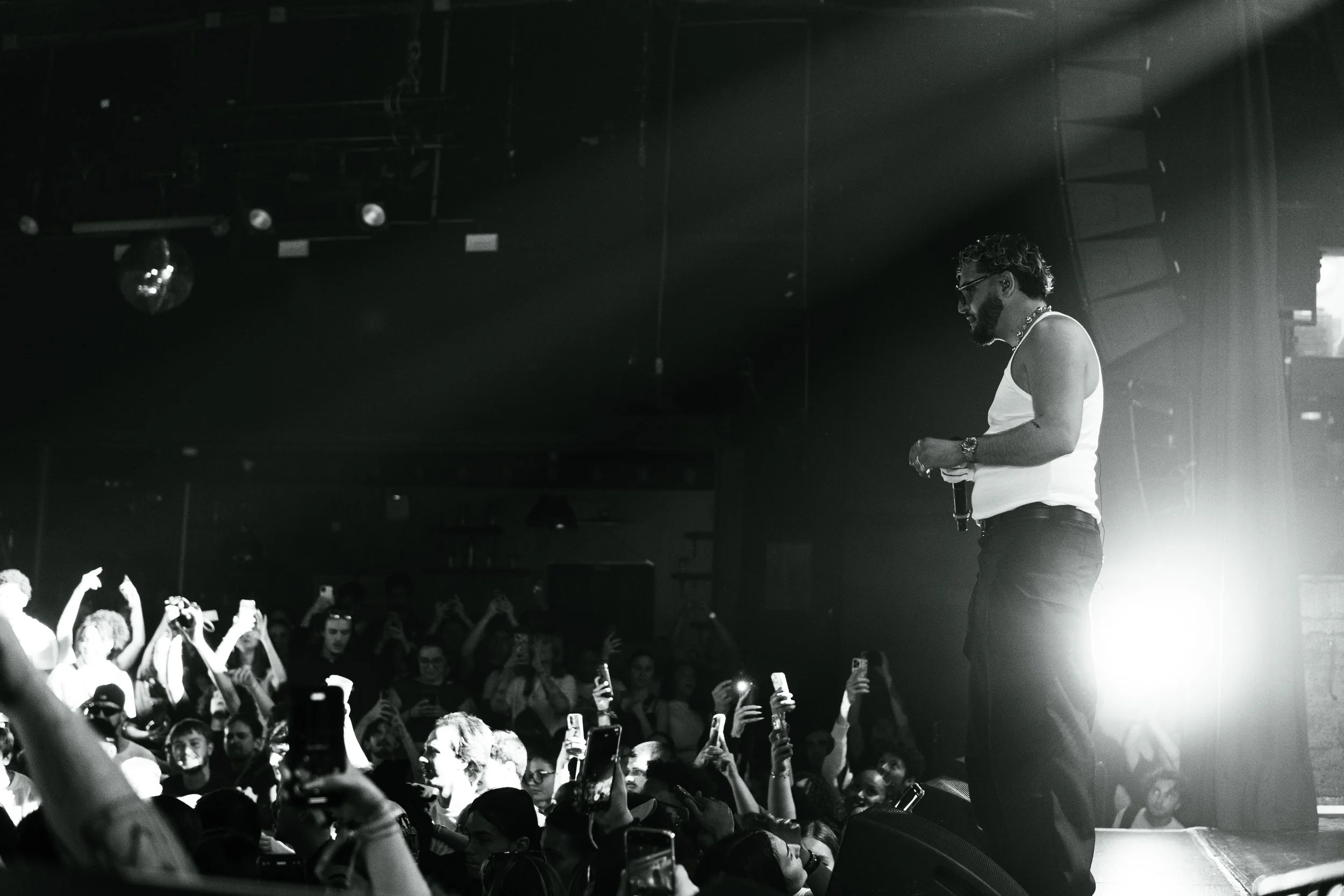 Un homme avec des lunettes, un t-shirt blanc sans manches et un collier, tenant un microphone, se tient sur scène face à une foule qui lève des téléphones et des appareils photo pour prendre des photos, dans une salle de concert ou un club. La scène 