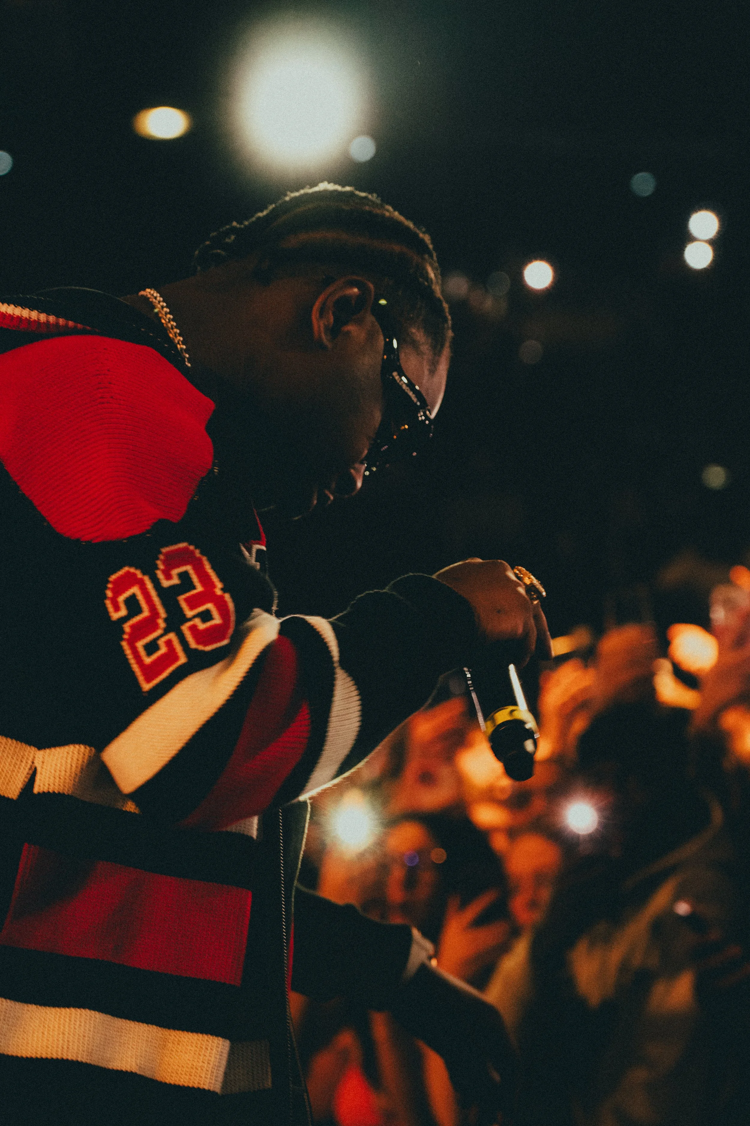 Une personne portant une veste à rayures rouges, blanches et noires, tenant un microphone, probablement lors d'une performance ou d'un discours, avec un public en arrière-plan.