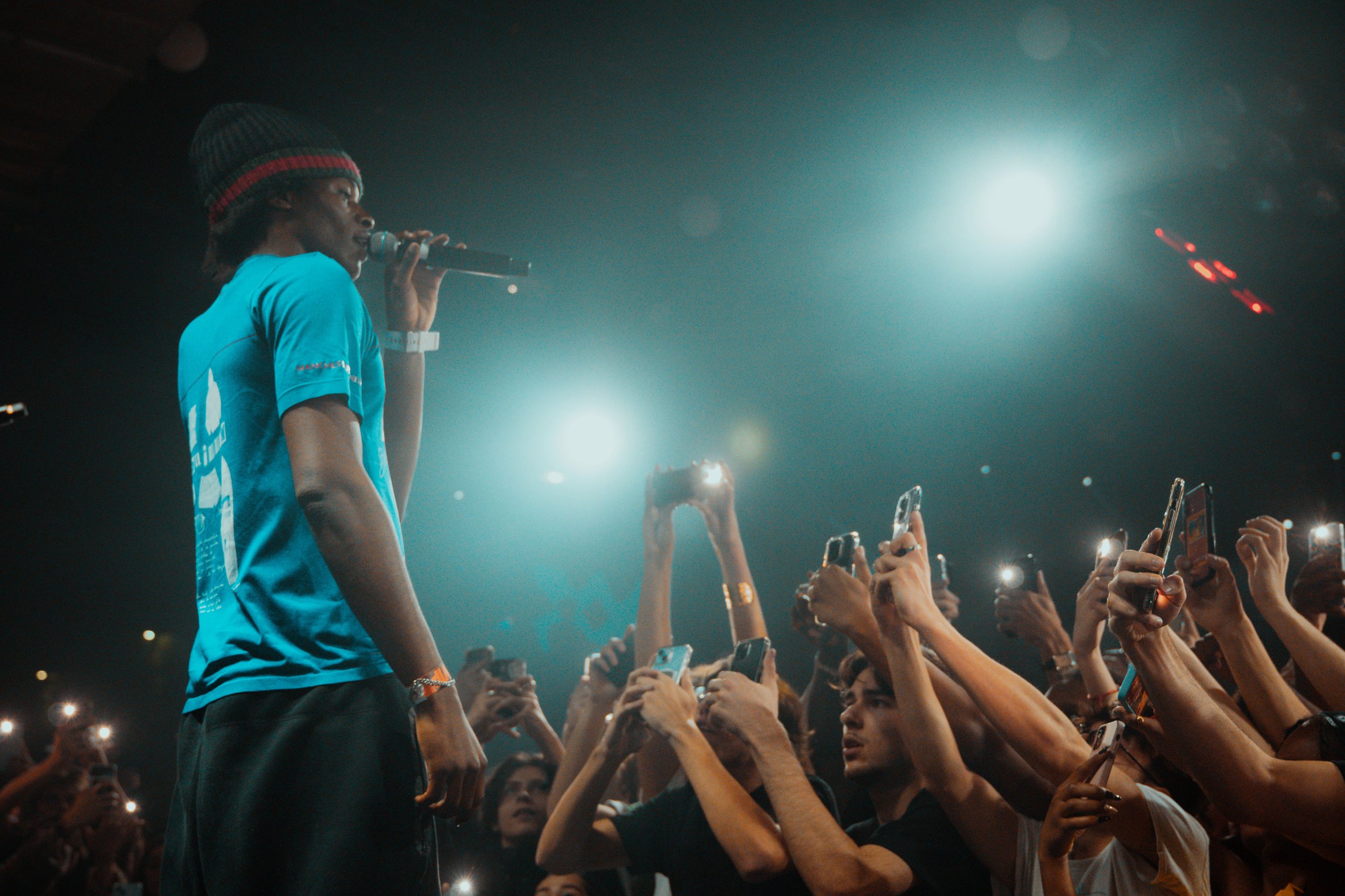 Un jeune homme en t-shirt bleu et bonnet noir avec rayures rouges chante dans un microphone lors d'un concert, entouré d'une foule de fans tenant des téléphones avec flashs allumés, sous un éclairage de scène.