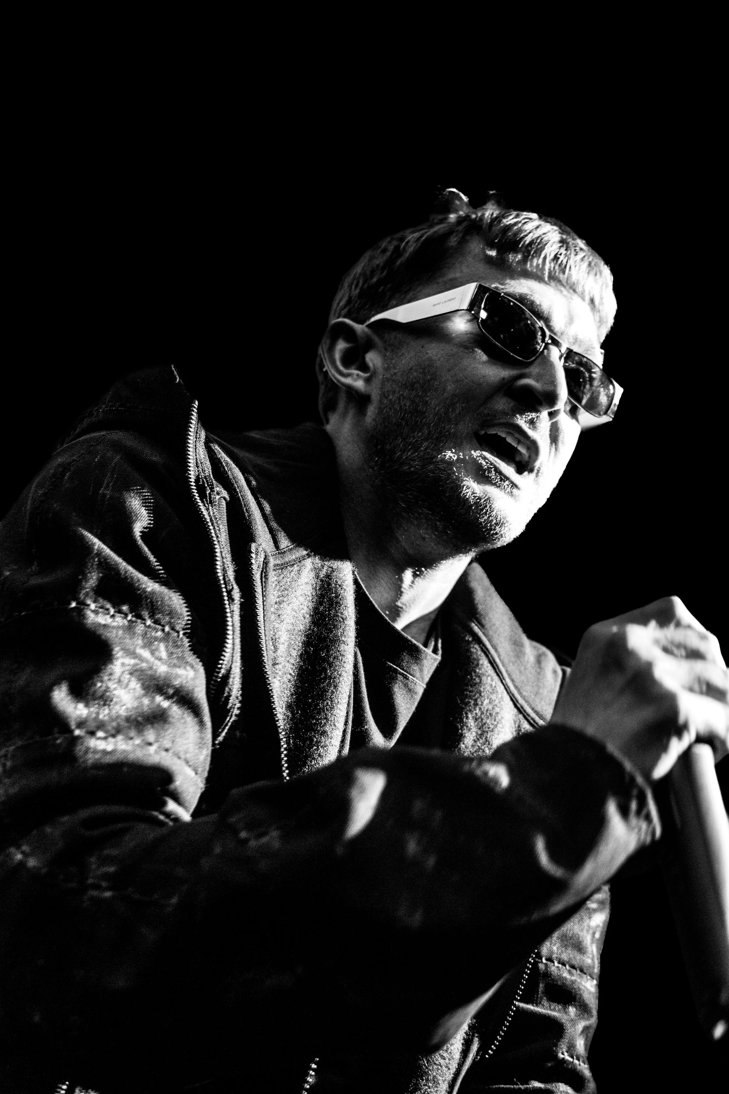 Un homme portant des lunettes de soleil, un blouson en cuir et une expression enthousiaste, parlant ou chantant dans un contexte sombre, en noir et blanc.