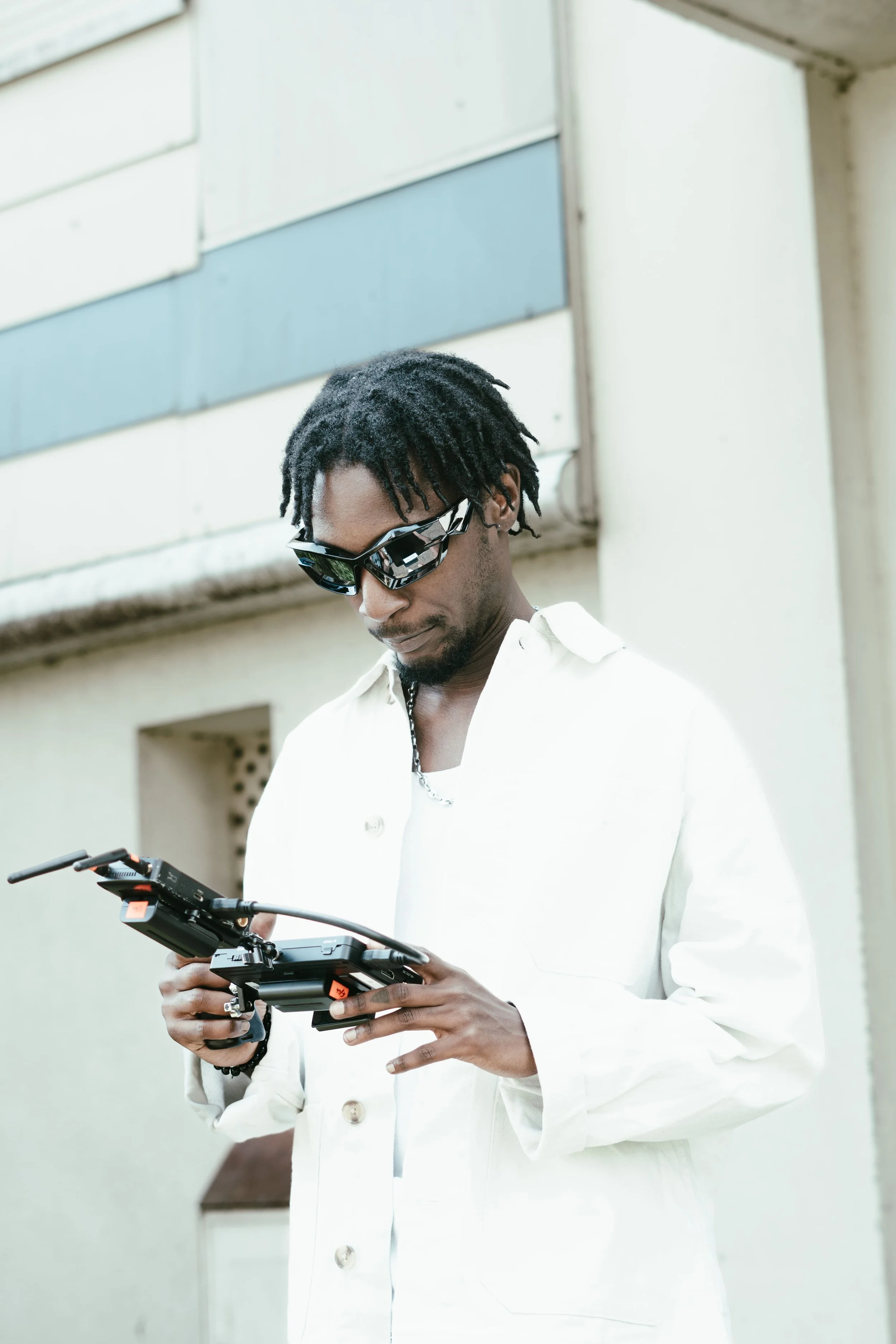 Un homme portant des lunettes de soleil et une chemise blanche tient une télécommande ou un contrôleur de drone, regardant l'appareil.