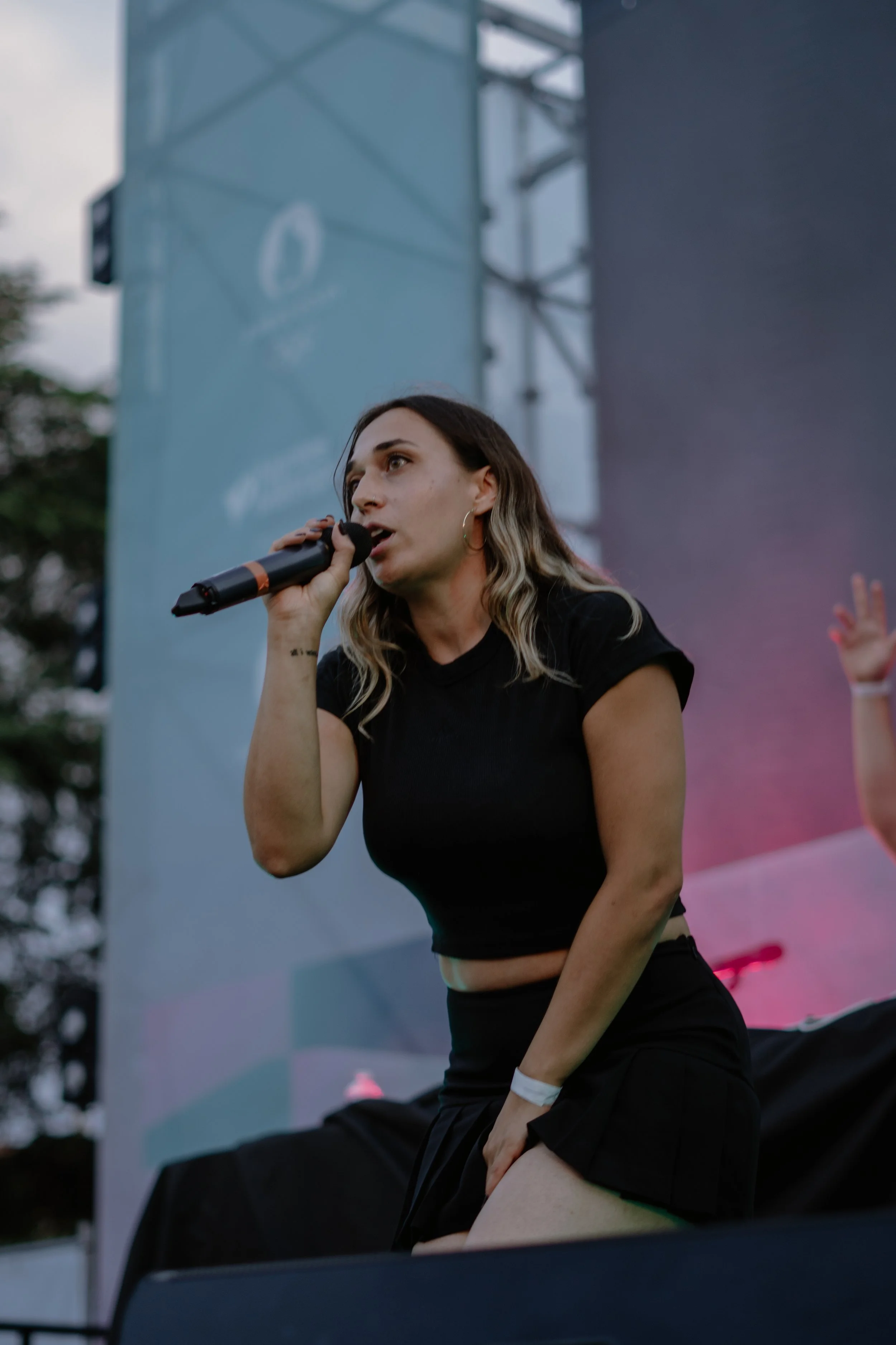 Une femme chante lors d'un concert en plein air, tenant un microphone, avec un arrière-plan de scène et d'arbres, semble en train de performer.
