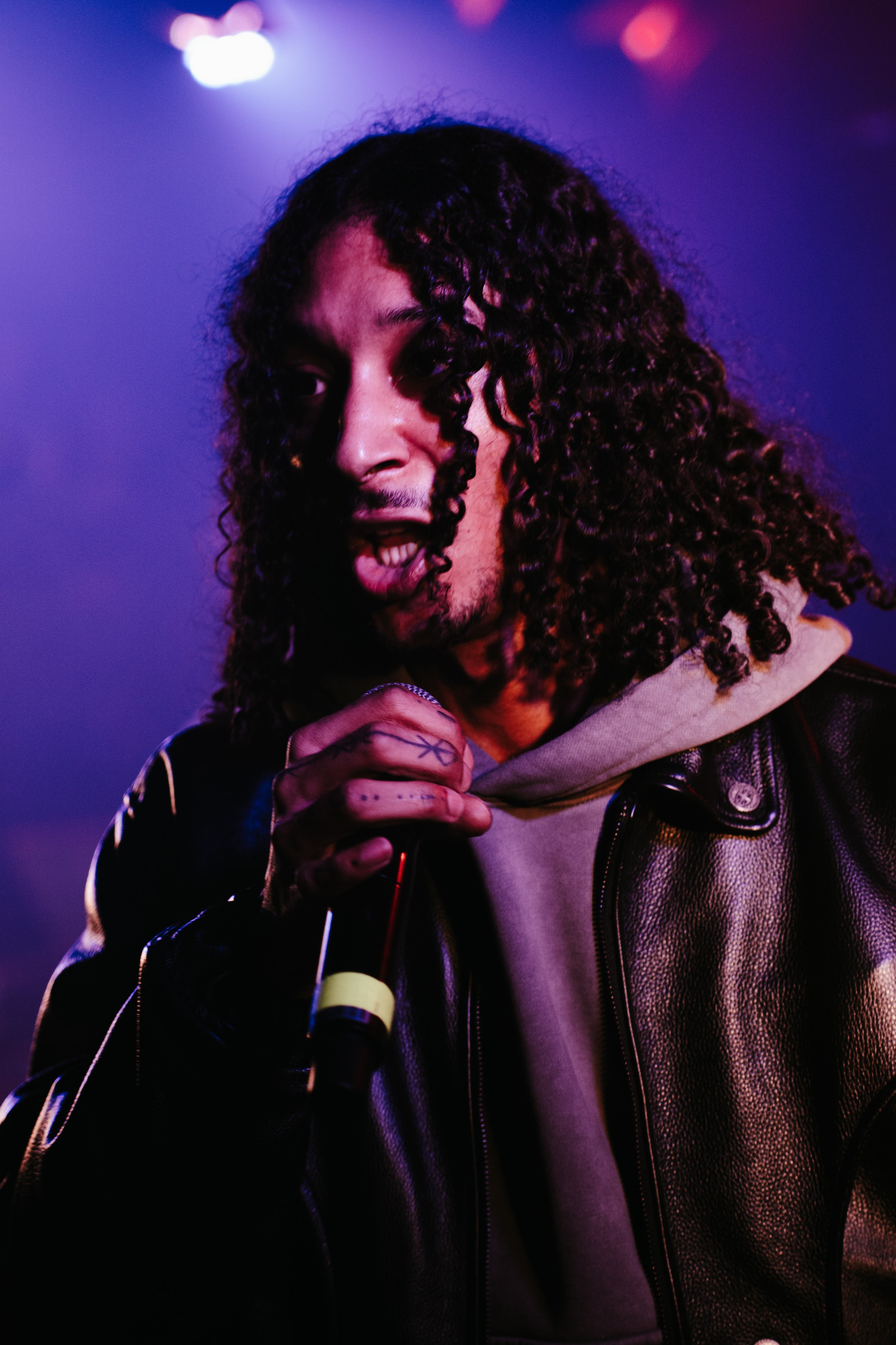 Un homme souriant, avec des cheveux bouclés, tenant un microphone, portant une veste en cuir et une capuche grise, dans un environnement éclairé violette et bleue.