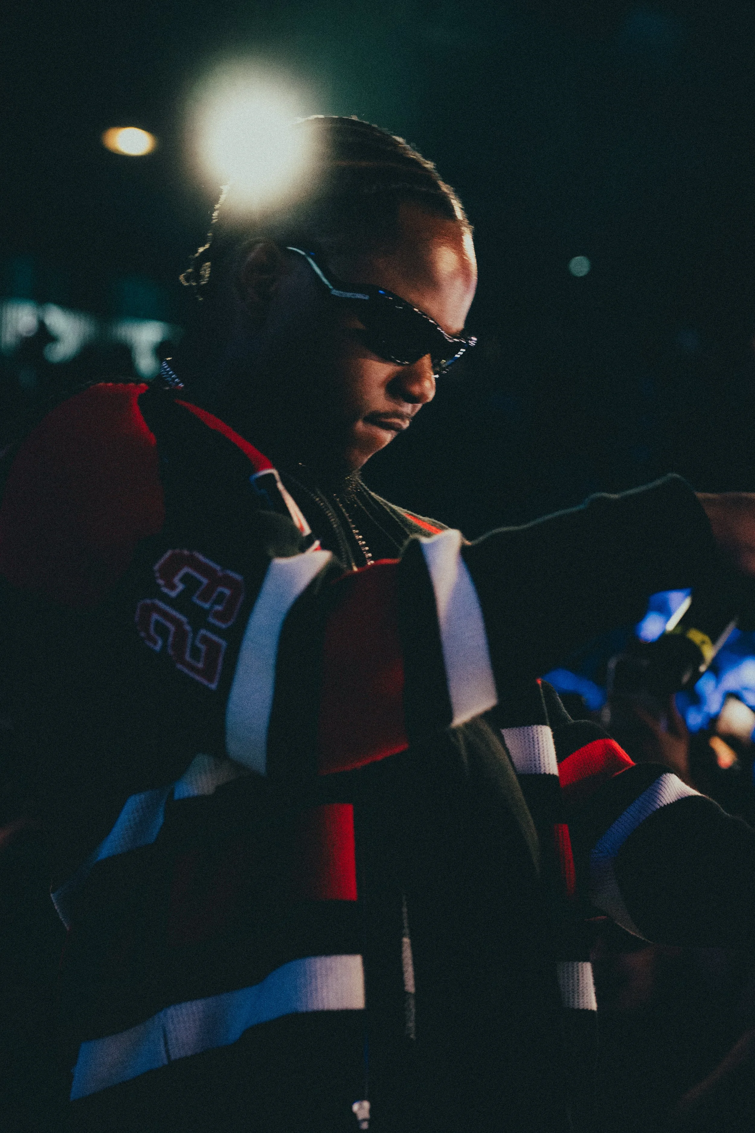 Un jeune homme portant des lunettes de soleil et une veste de hockey aux couleurs rouge, noir, blanc et bleu, dans un environnement sombre avec un éclairage dramatique.