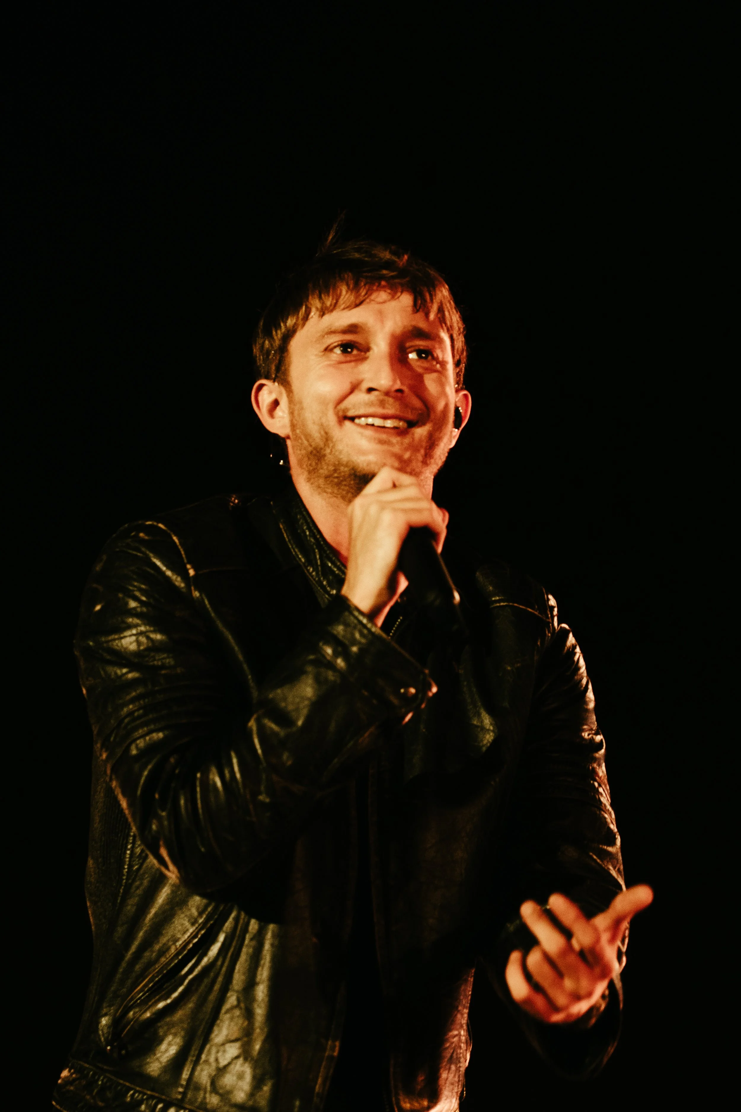 Homme souriant portant un blouson en cuir noir, parlant ou chantant lors d'une performance sur scène.