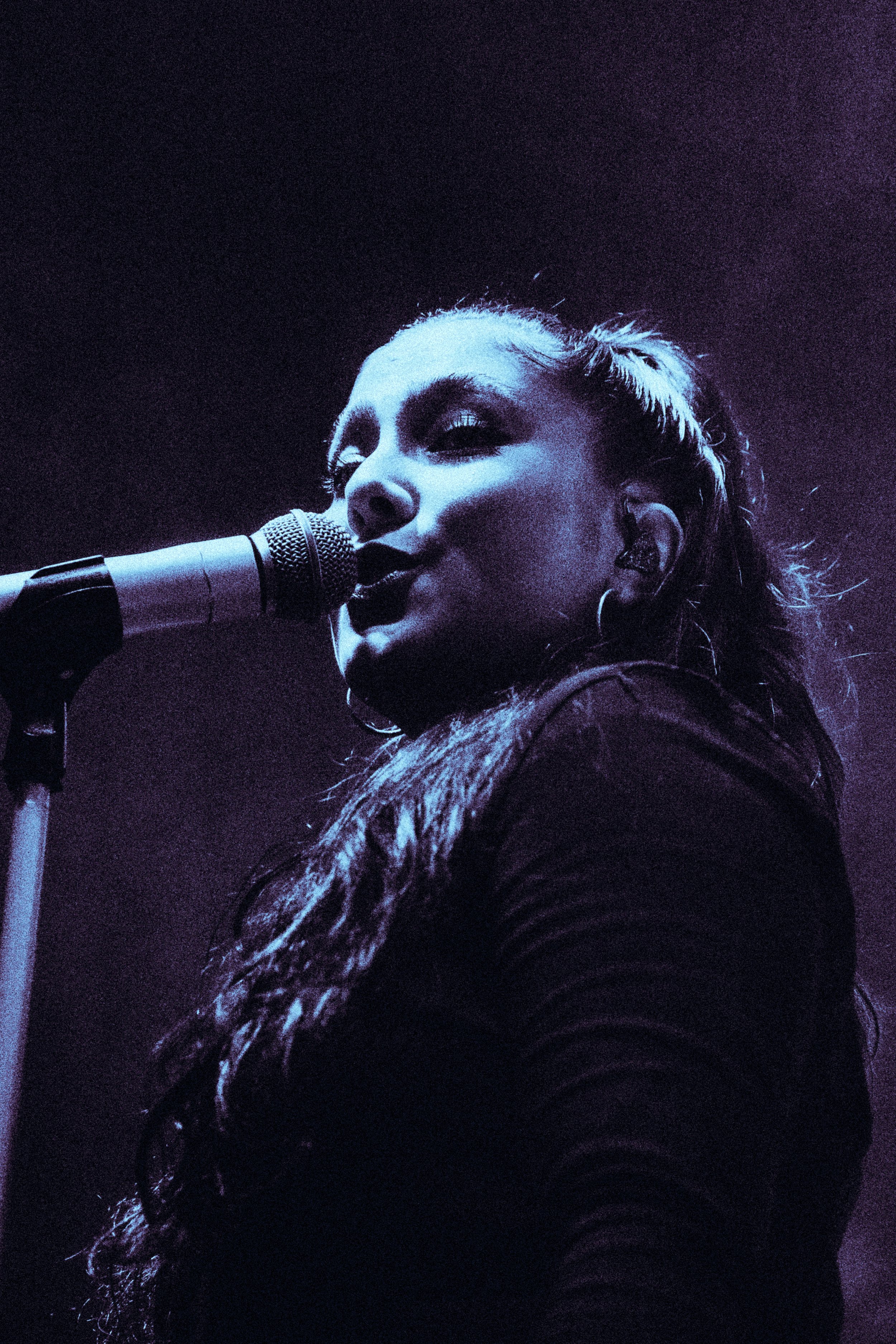 Une femme chante dans un microphone, éclairée par une lumière violette, avec une expression concentrée.