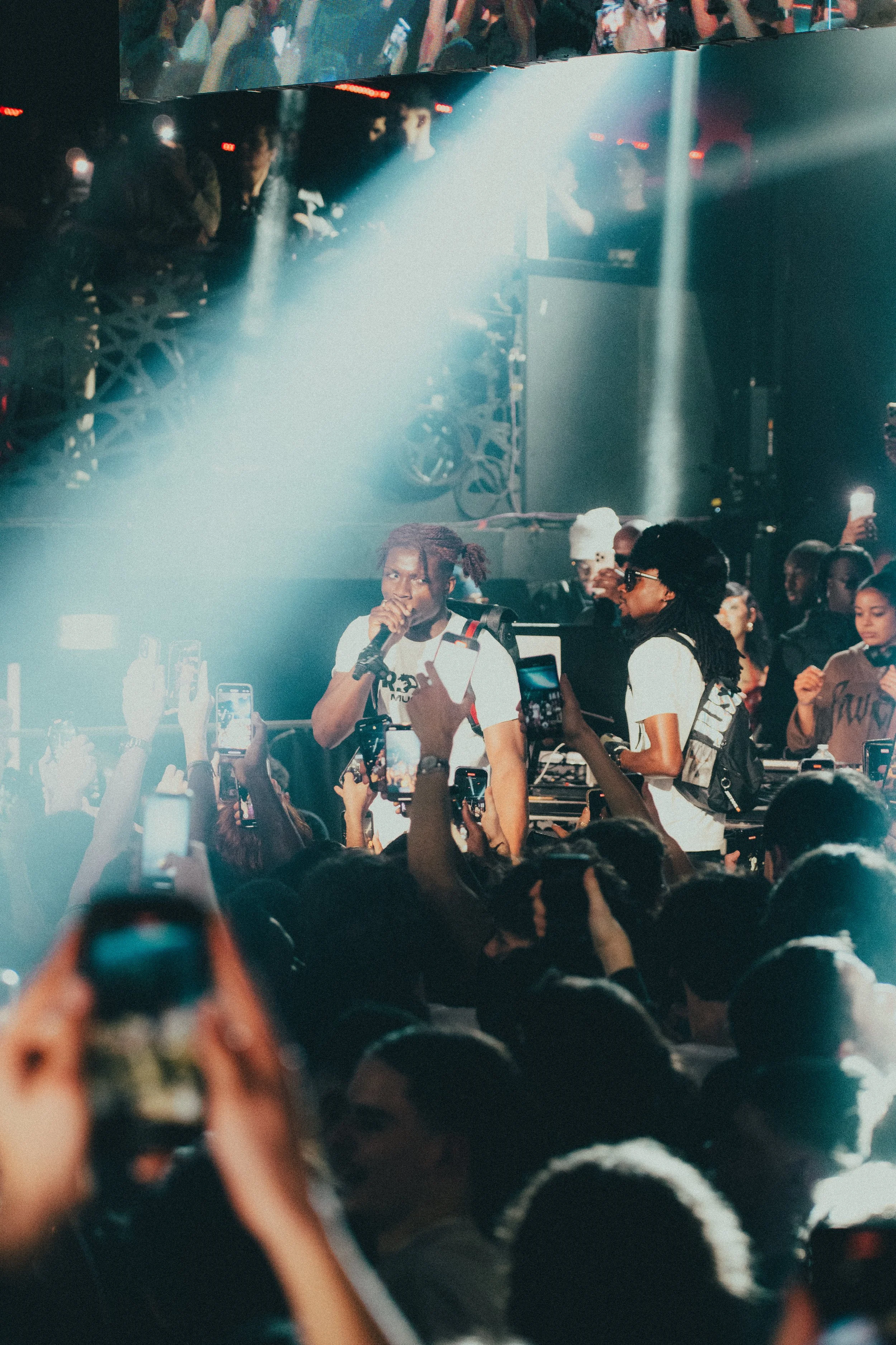 Un artiste rap en train de chanter sur scène lors d'un concert bondé, entouré de fans qui prennent des photos et des vidéos.