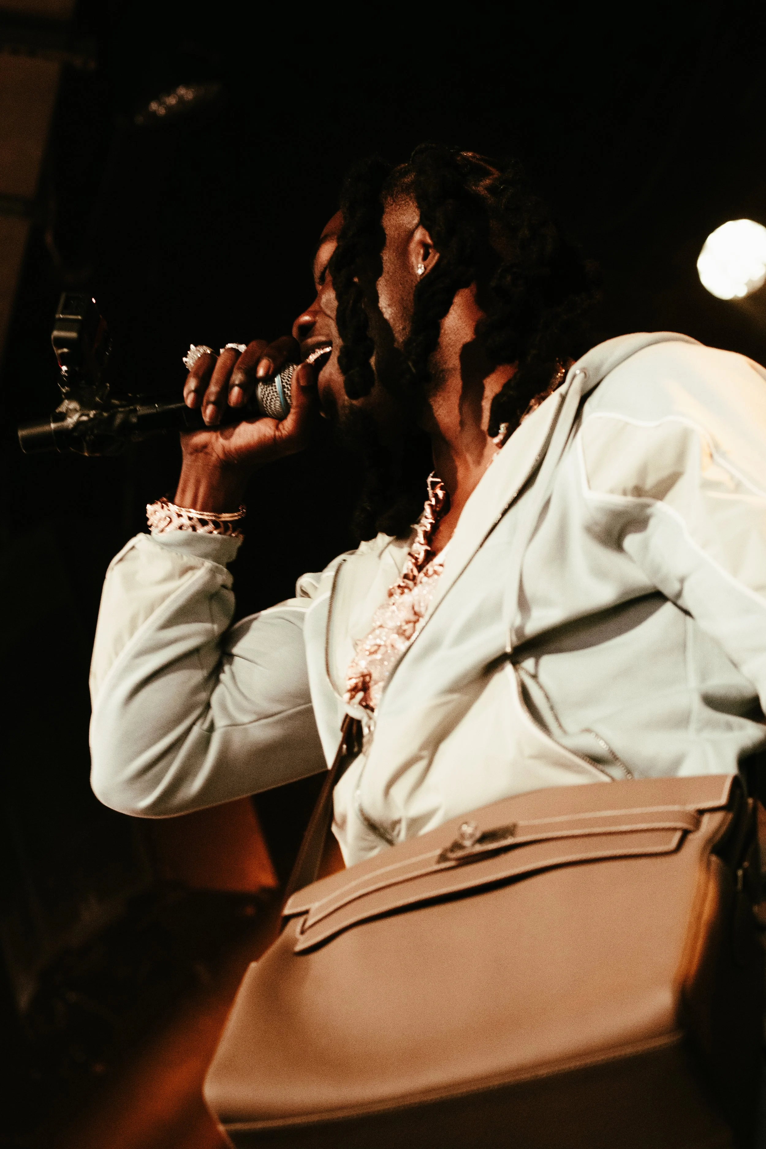 Une femme en train de chanter ou parler dans un microphone lors d'une performance, portant un vêtement blanc, des bijoux, et un sac beige.
