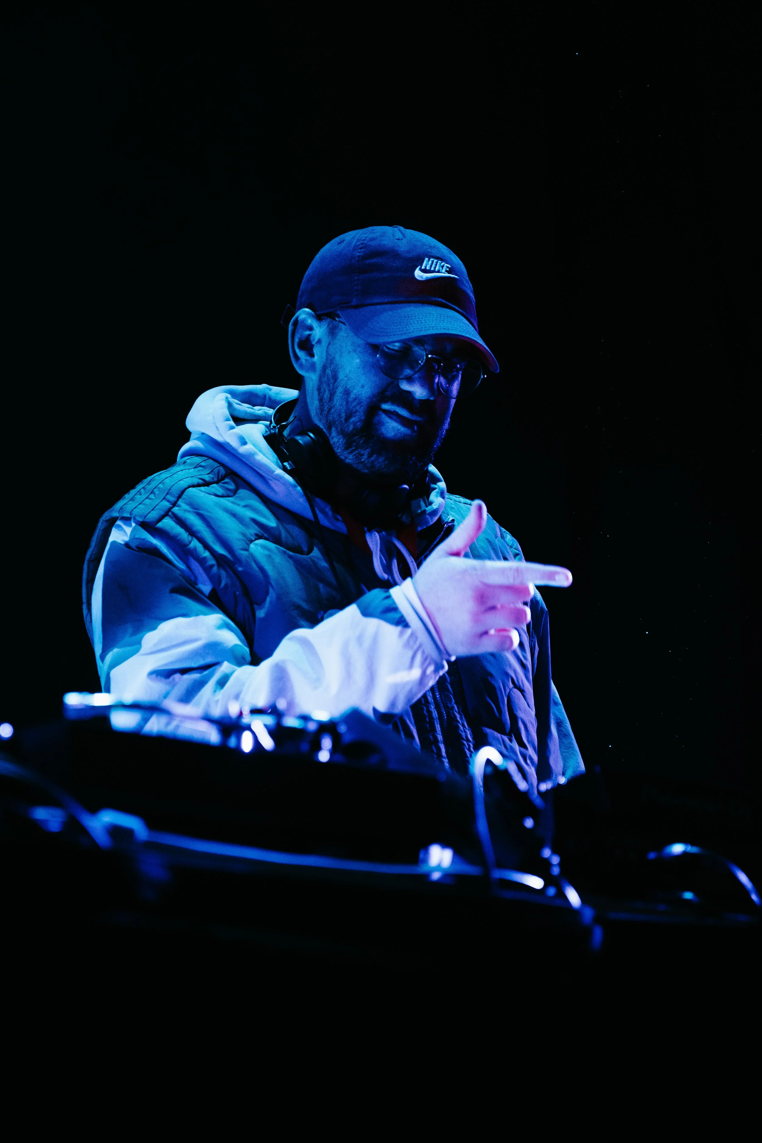 DJ avec casque et lunettes, pointant avec sa main, éclairé par des lumières bleues, sur une scène sombre.