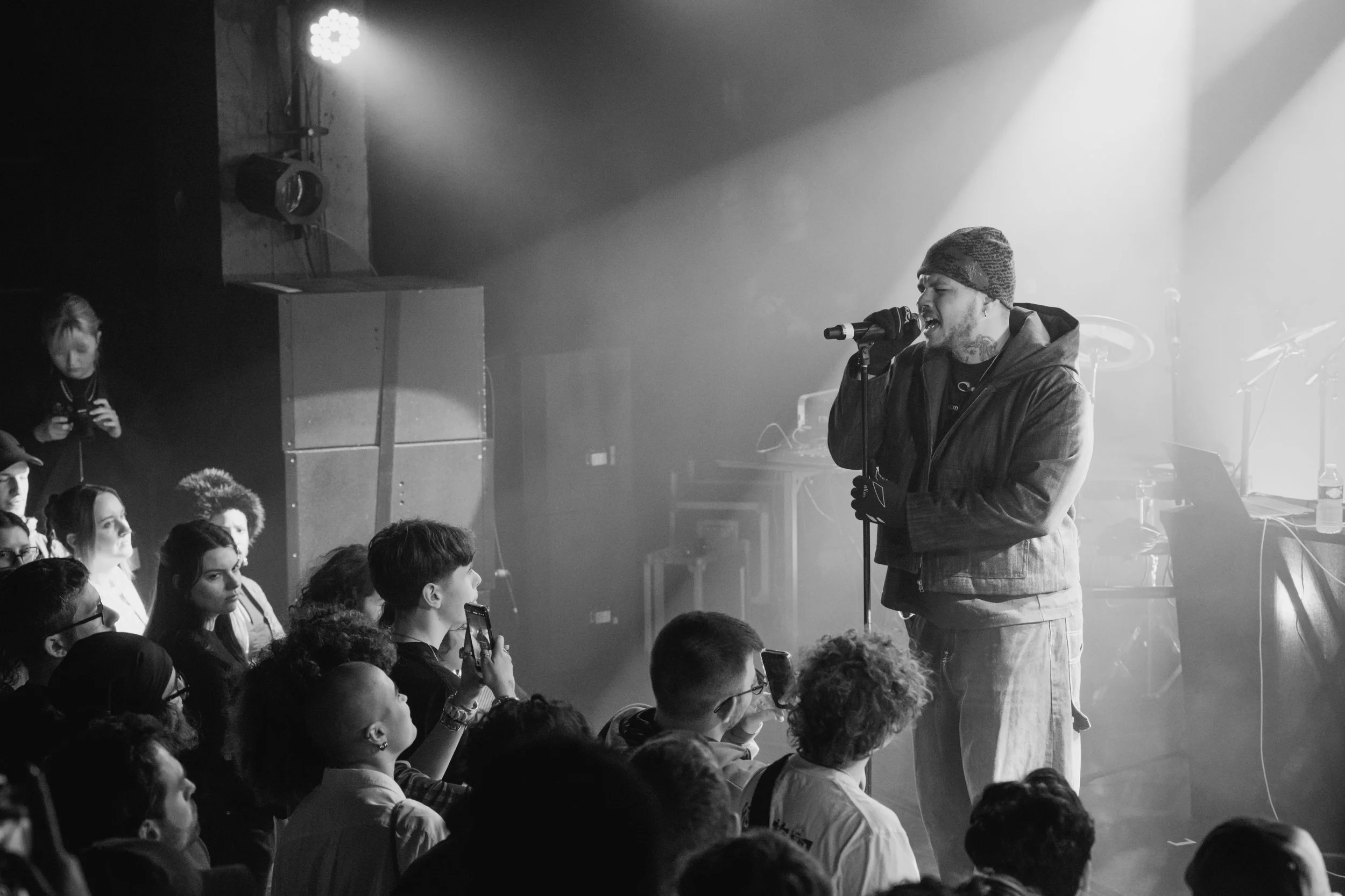 Un homme chante dans un microphone sur scène devant un public. L'ambiance est sombre avec des lumières tamisées.