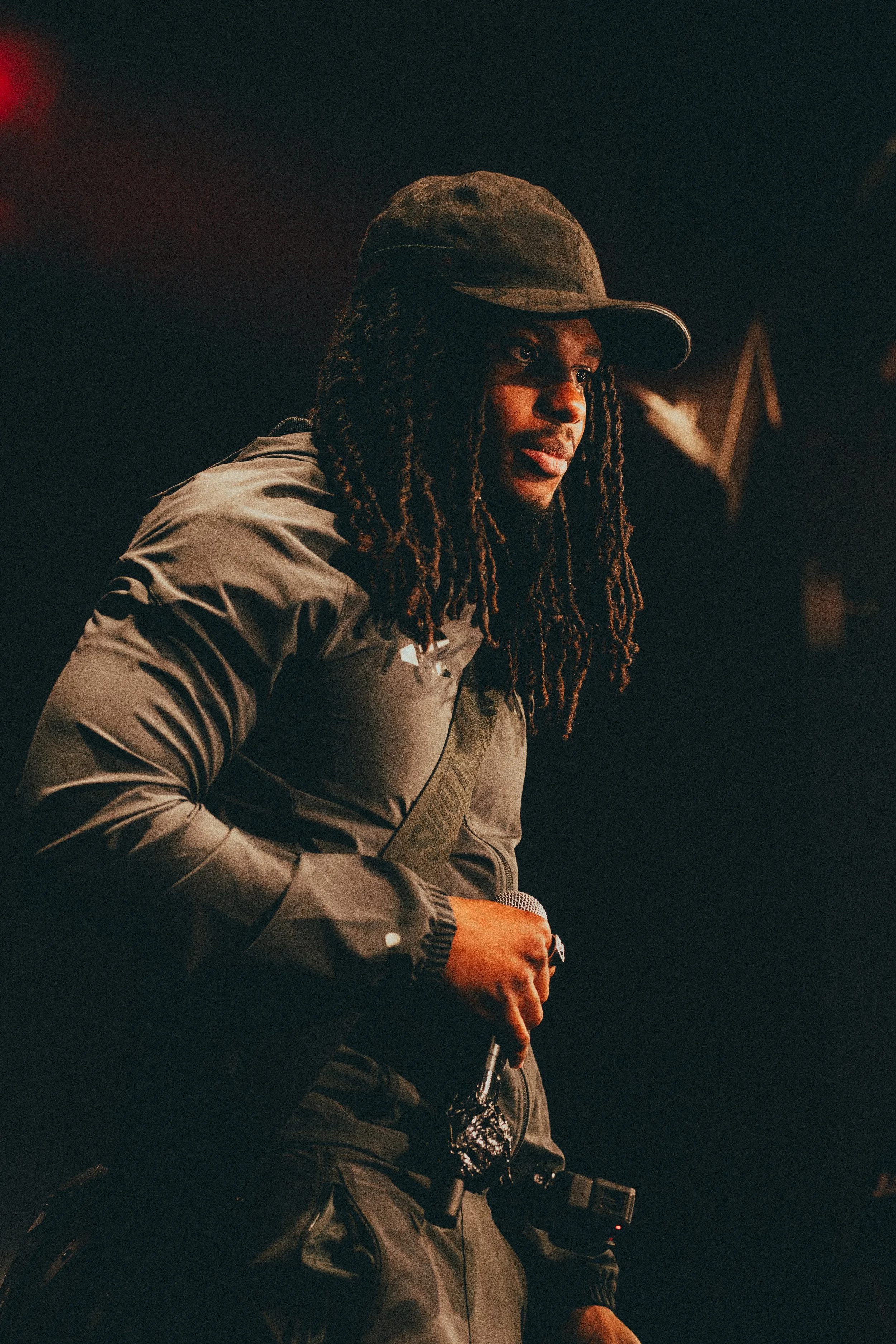 Une personne avec des dreadlocks portant une casquette et une veste noire tient un microphone dans une main dans un environnement sombre.