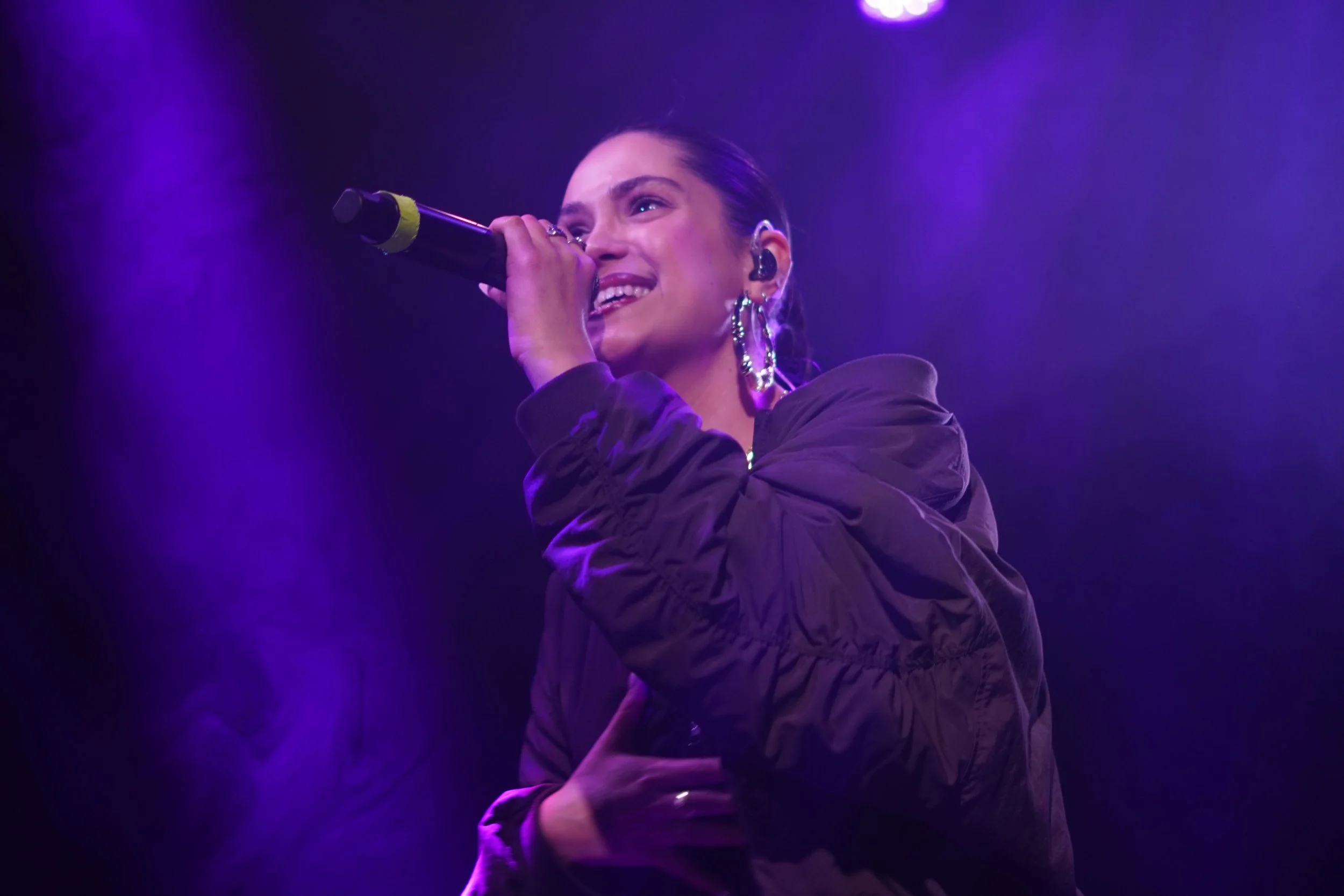 Une femme chante dans un micro, éclairée par des lumières violettes, portant des boucles d'oreilles pendantes et une veste sombre, en pleine performance sur scène.