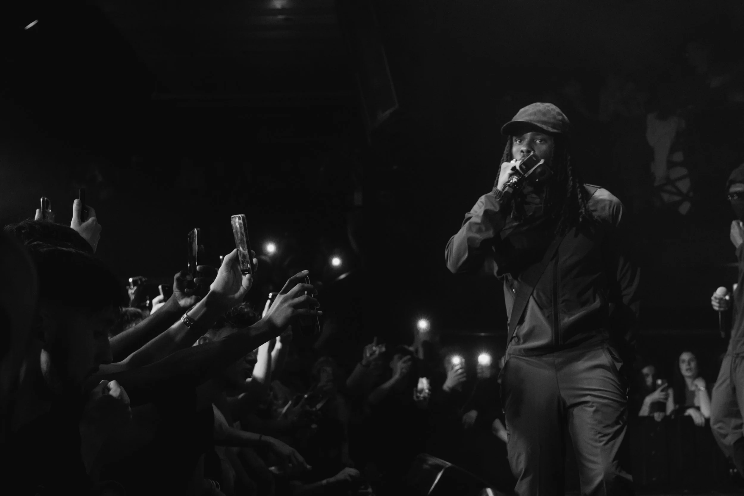 Un artiste de rap sur scène lors d'un concert, entouré par un public qui l'enregistre avec leurs téléphones, en noir et blanc.