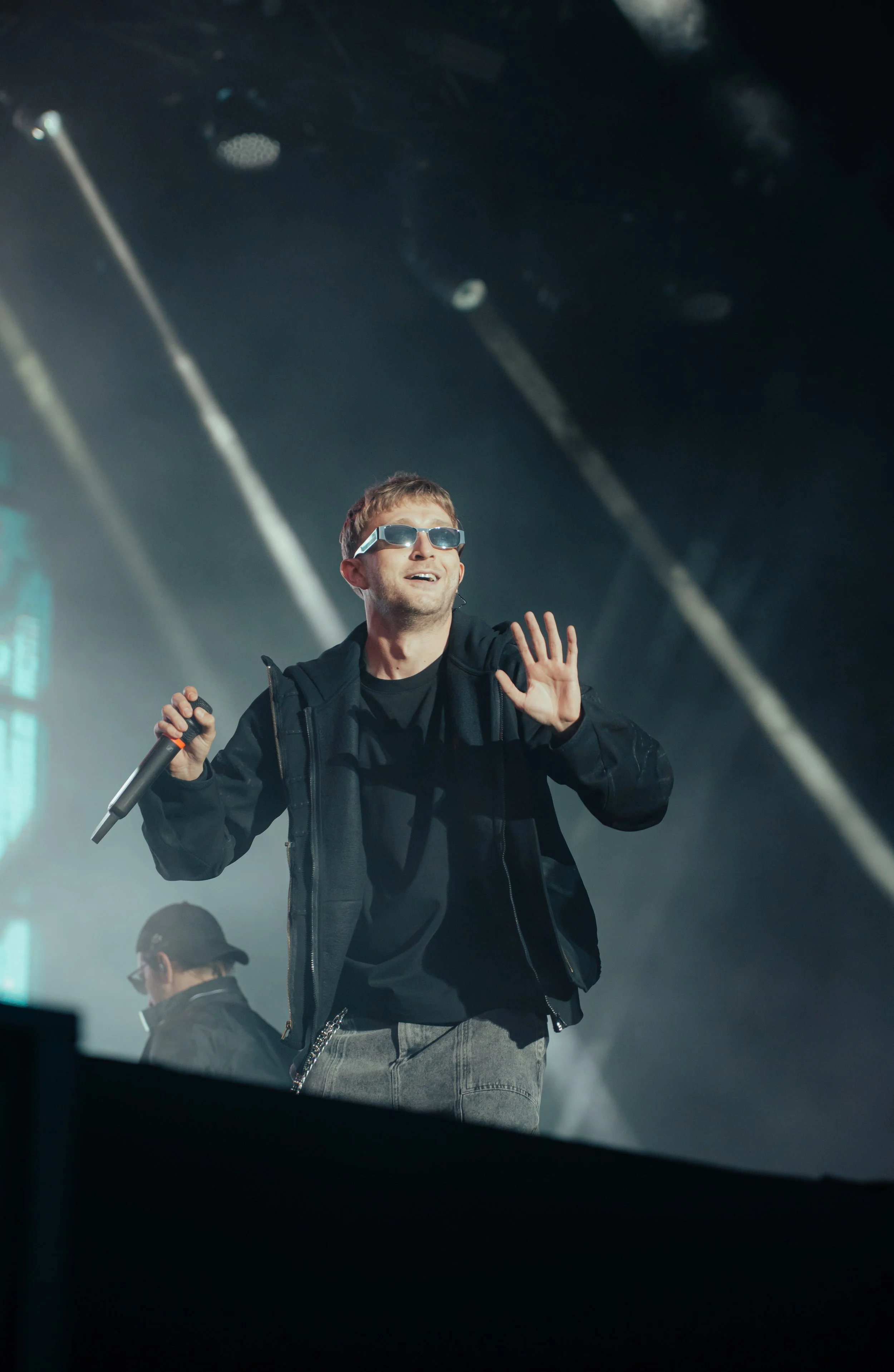Un homme portant des lunettes de soleil, en tenue noire, souriant et faisant signe de la main lors d'une performance sur scène