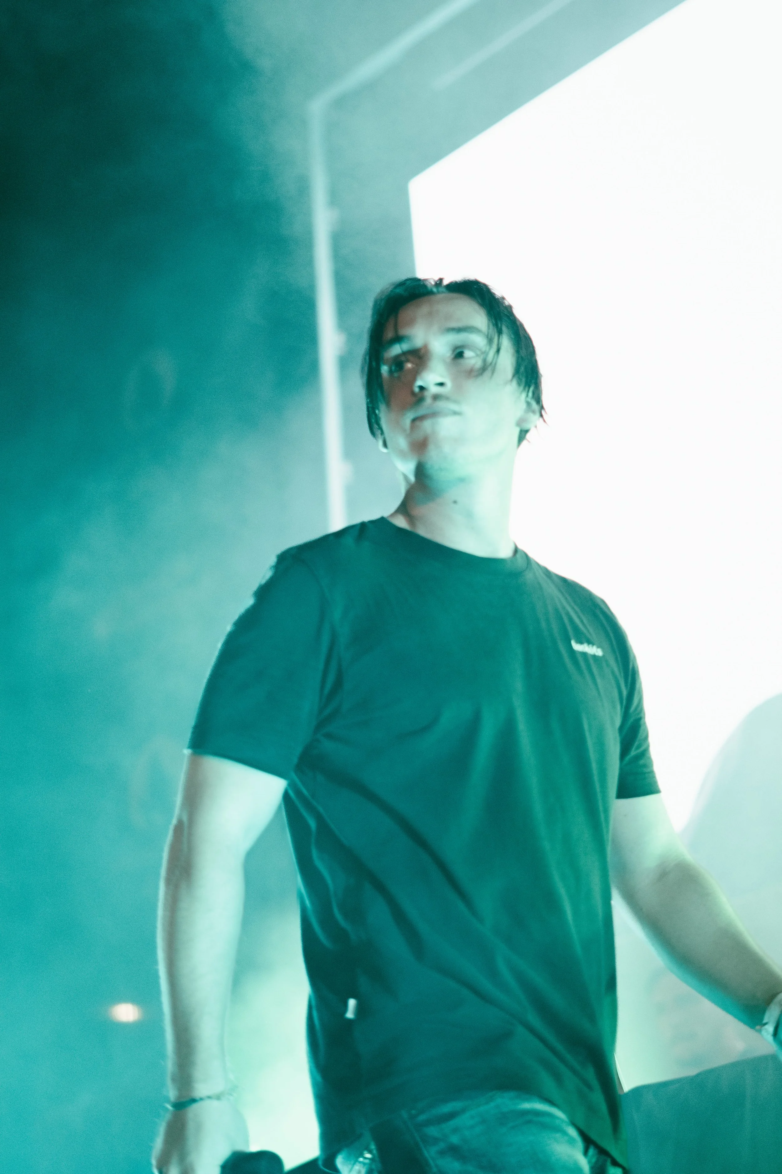 Un jeune homme avec des cheveux noirs et une expression sérieuse, portant un t-shirt noir, se tenant dans une ambiance sombre avec un éclairage vert et blanc