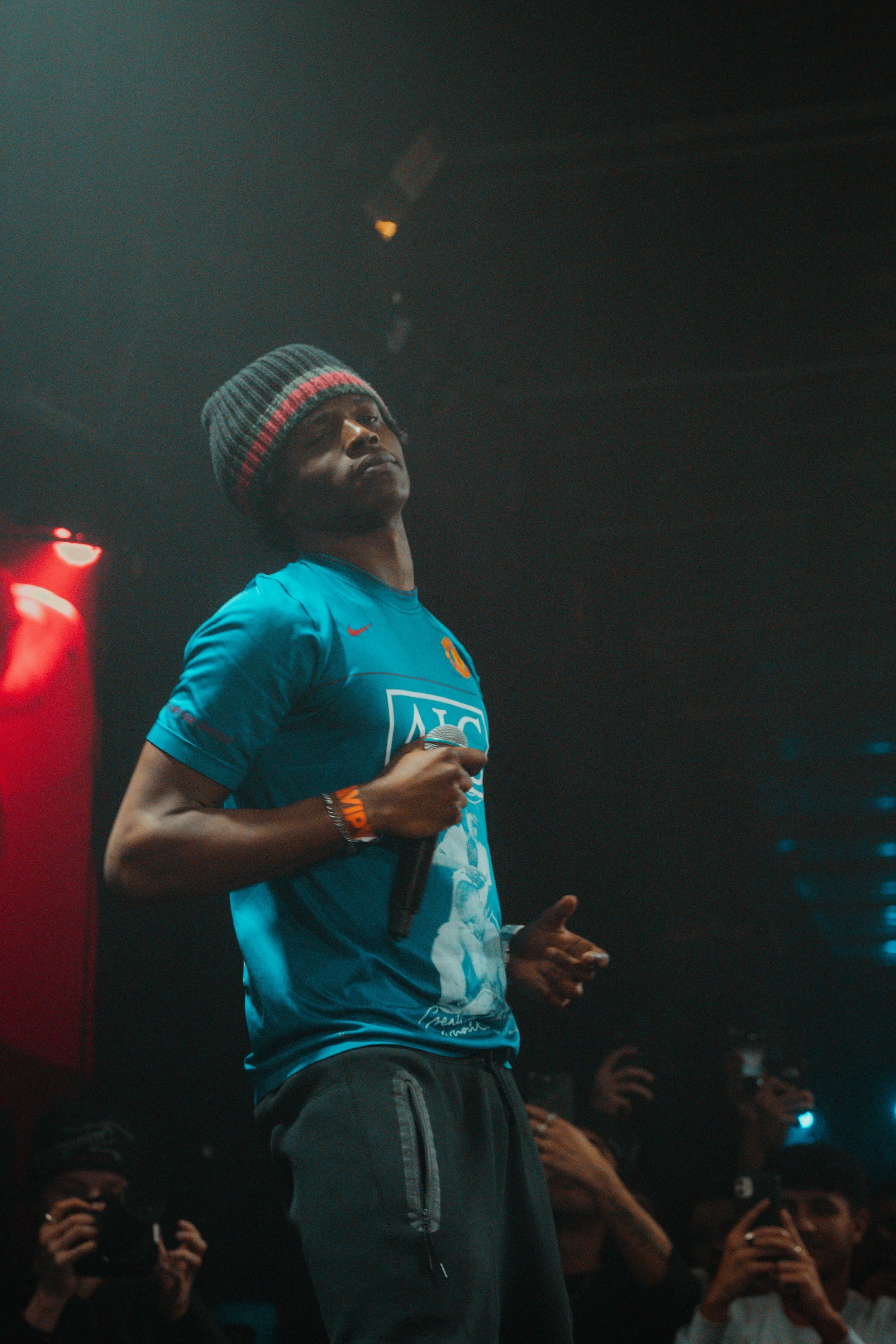 Un homme avec un bonnet gris et une t-shirt bleu, tenant un microphone, dans une ambiance sombre, entouré de personnes prenant des photos.