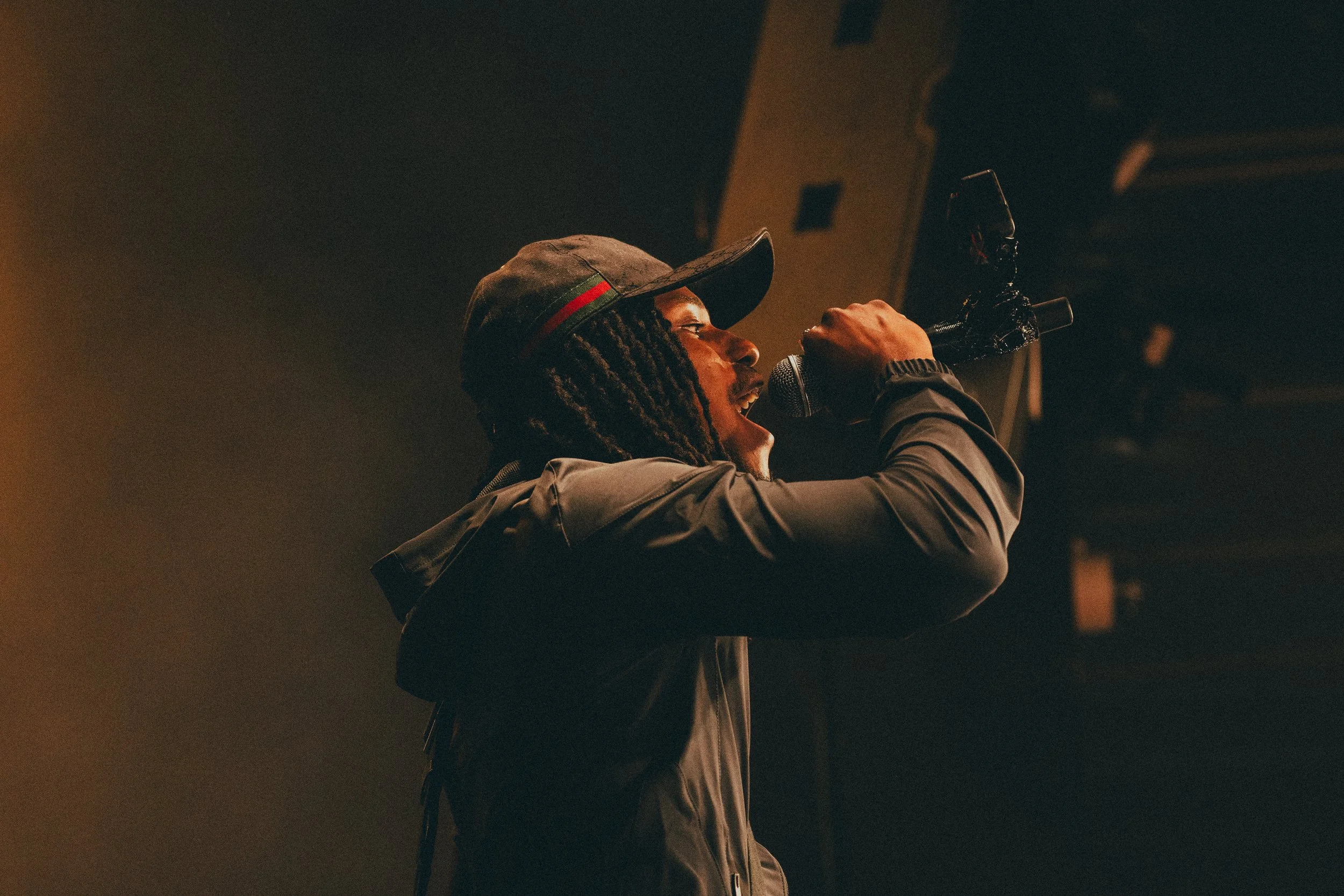 Un homme souriant, portant une casquette, chantant dans un microphone dans une scène sombre.
