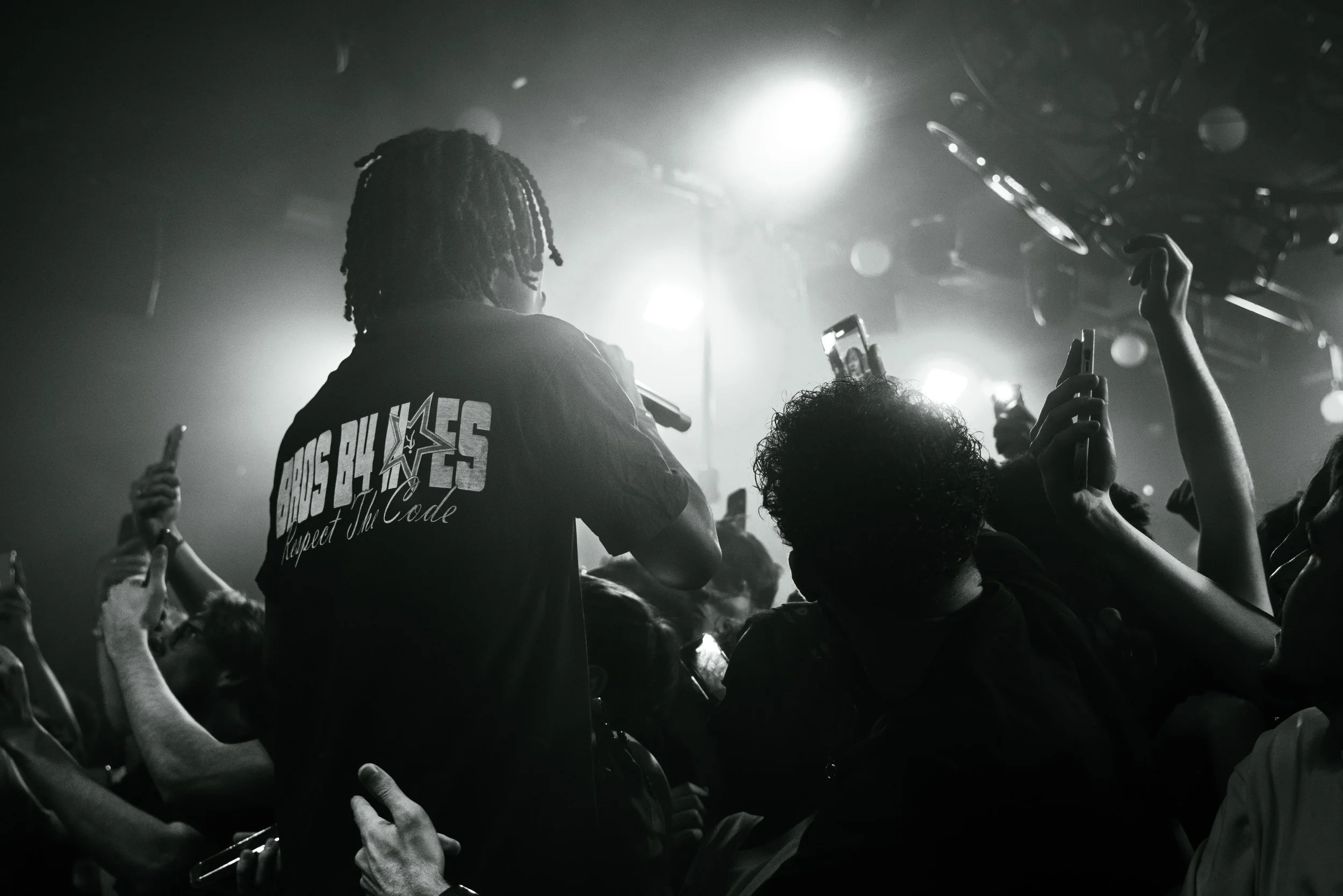 Une personne avec des dreadlocks porte un t-shirt avec le texte "CROSS BLONDES Respect The Code" lors d'un concert ou événement musical, entourée d'une foule de spectateurs prenant des photos avec leurs téléphones, dans un environnement sombre avec d