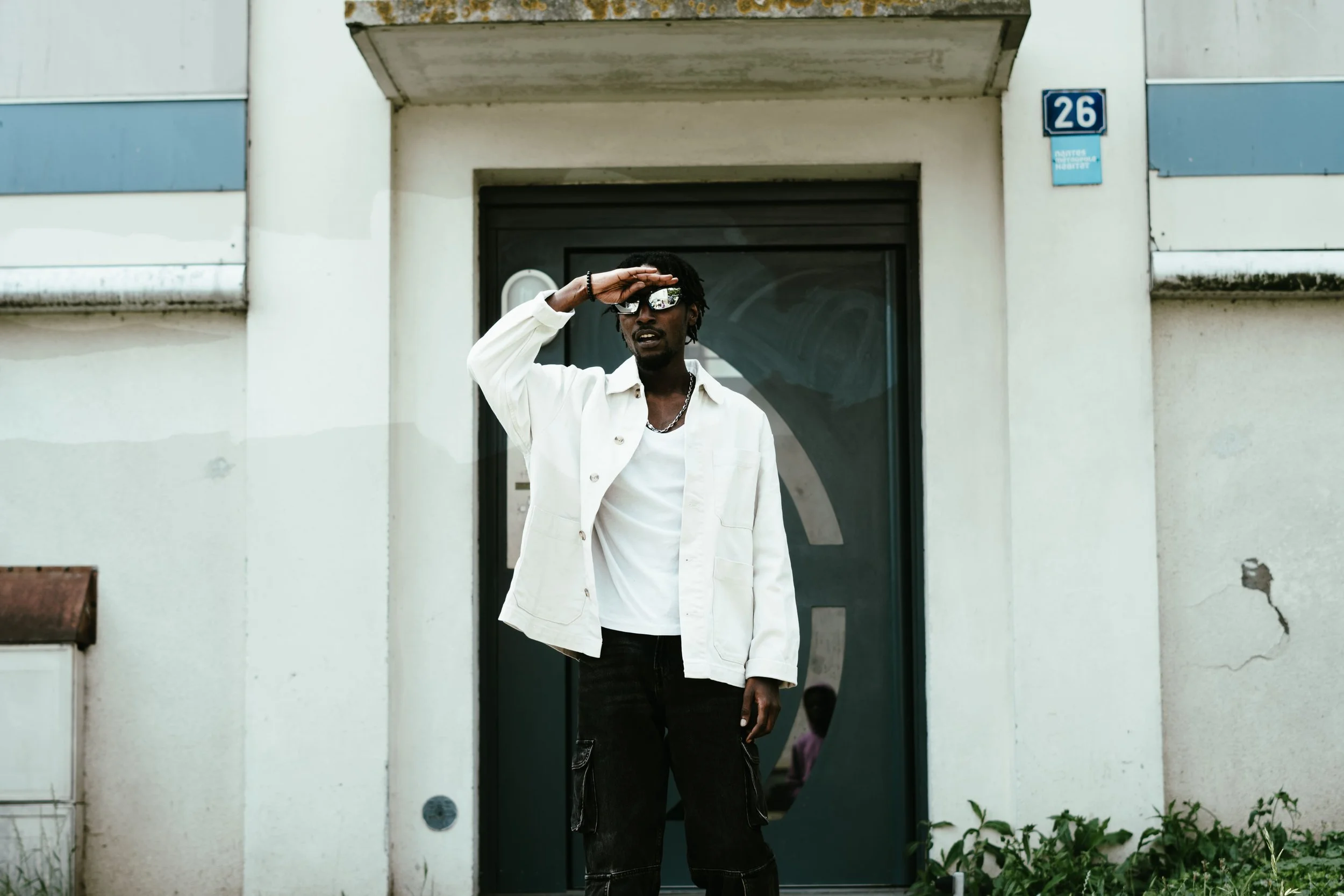 Un homme portant des lunettes de soleil, une veste blanche, une chaîne, et un t-shirt blanc, se tient devant une porte noire dans une rue urbaine, faisant un geste de salutation ou de recherche du soleil avec sa main.