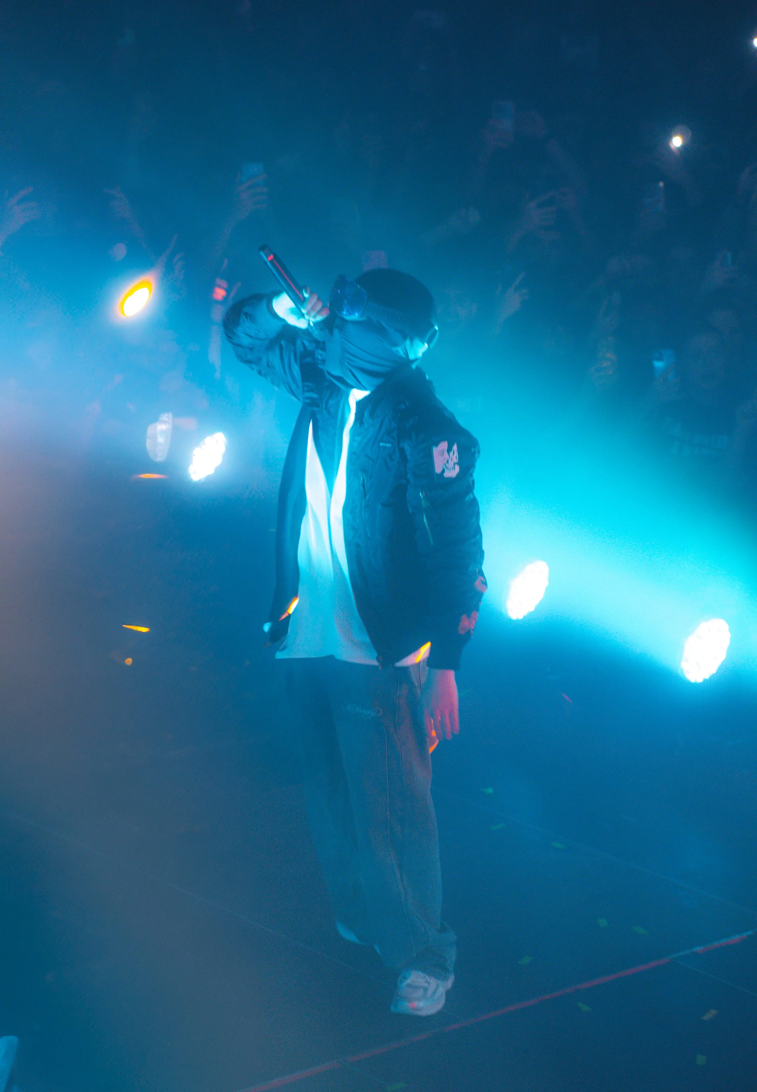 Un artiste performe sur scène, portant une veste noire avec une capuche, devant un fond de lumière bleue et blanche, entouré par une foule de spectateurs.