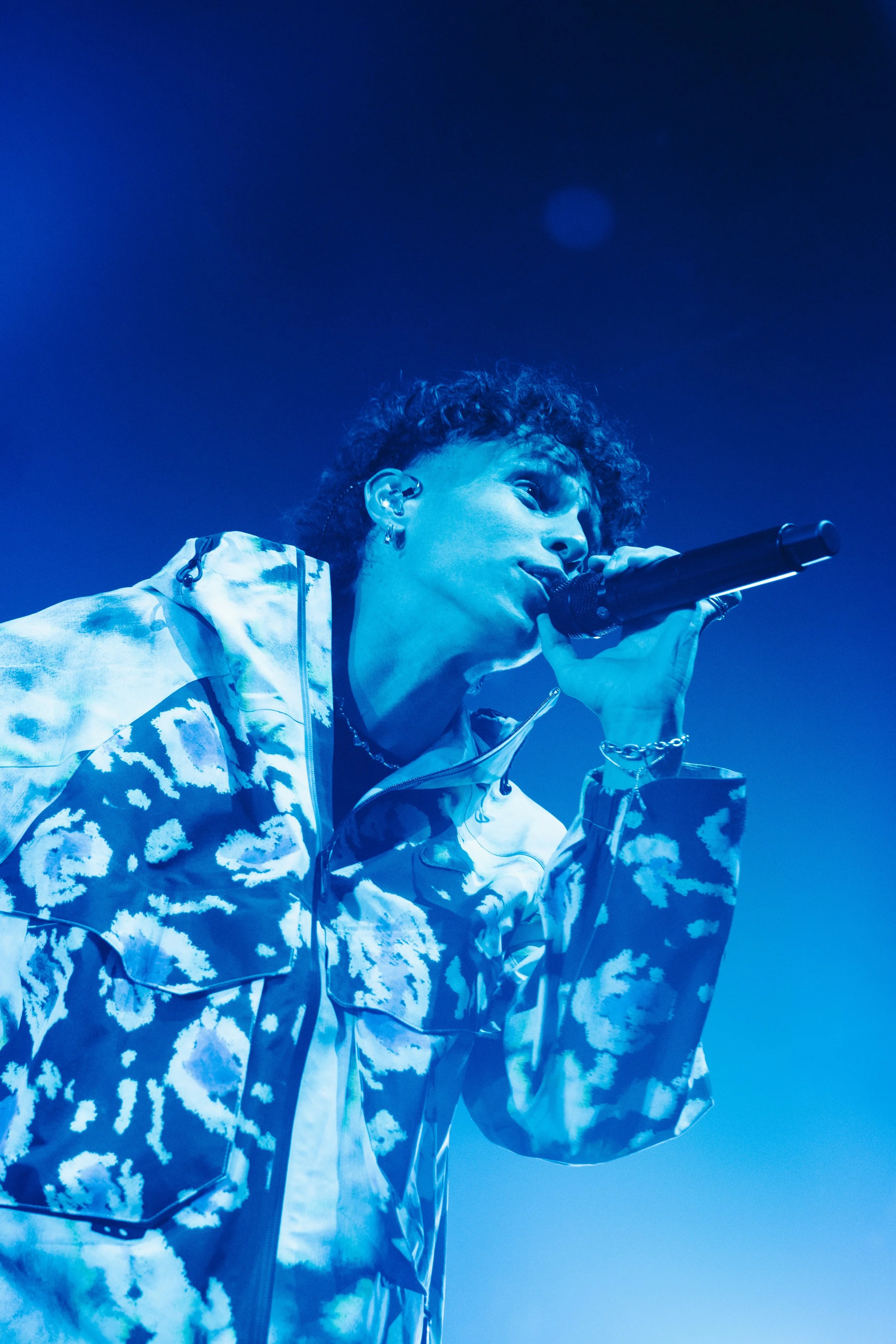 Un artiste en train de chanter avec un microphone sur scène, vêtue d'une veste à motif nuage en bleu et blanc et éclairée en bleuté.