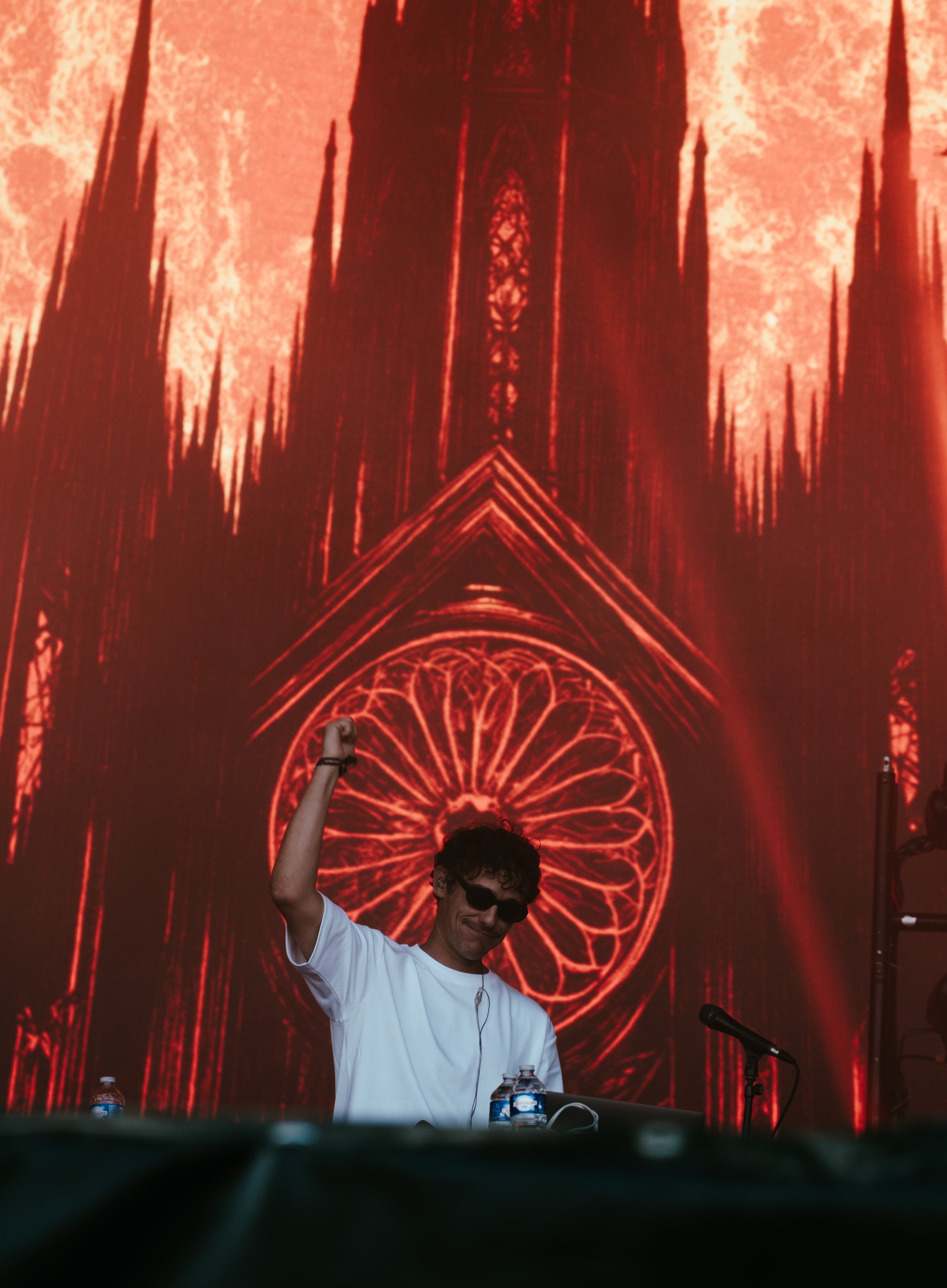 Un DJ avec des lunettes de soleil, chantant ou mimant un geste, devant un grand écran avec une image rouge d'une cathédrale gothique, probablement Notre-Dame.