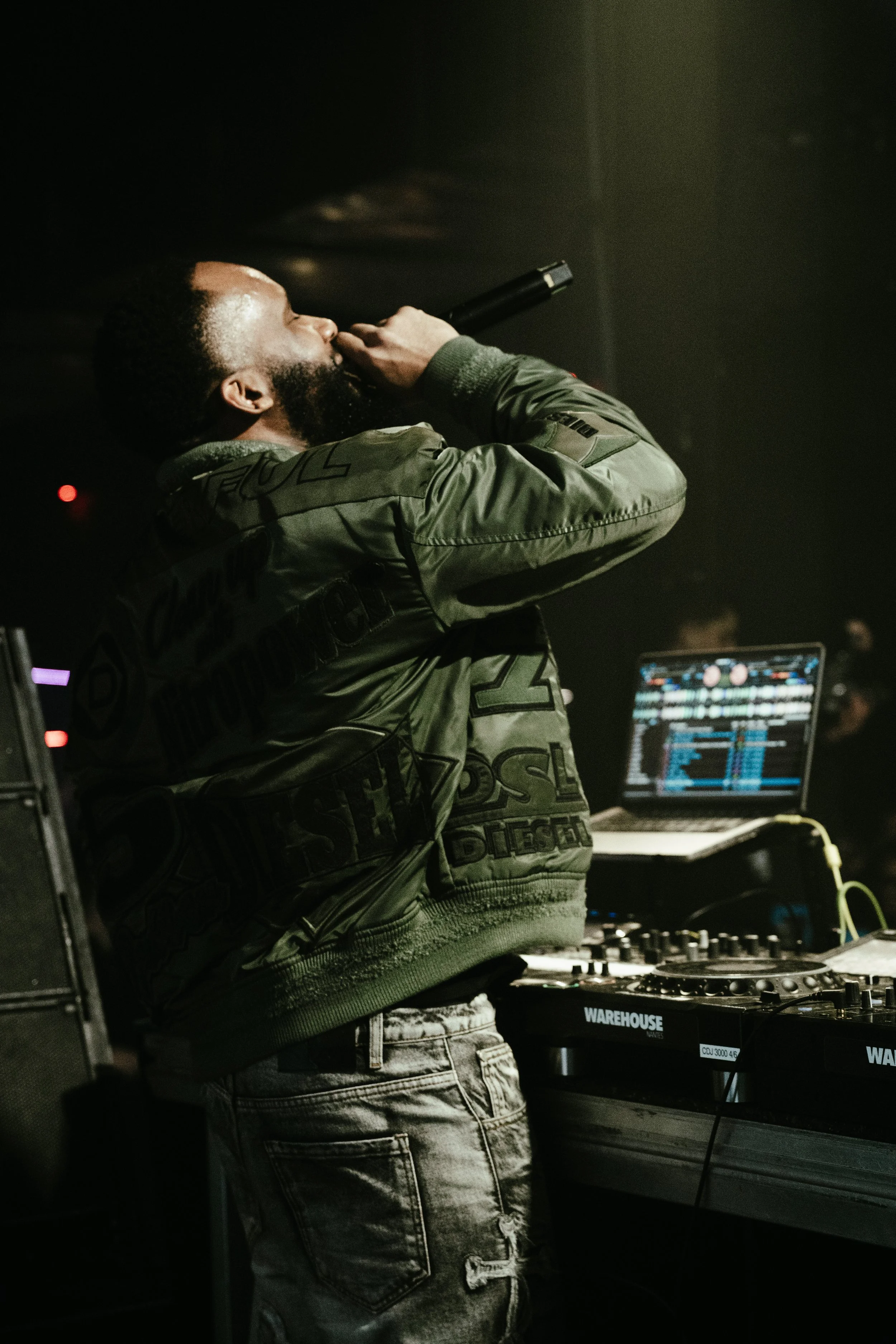 Un DJ en train de mixer dans une ambiance sombre, portant une veste olive avec des inscriptions, à côté d'un ordinateur portable et d'une console de mixage.