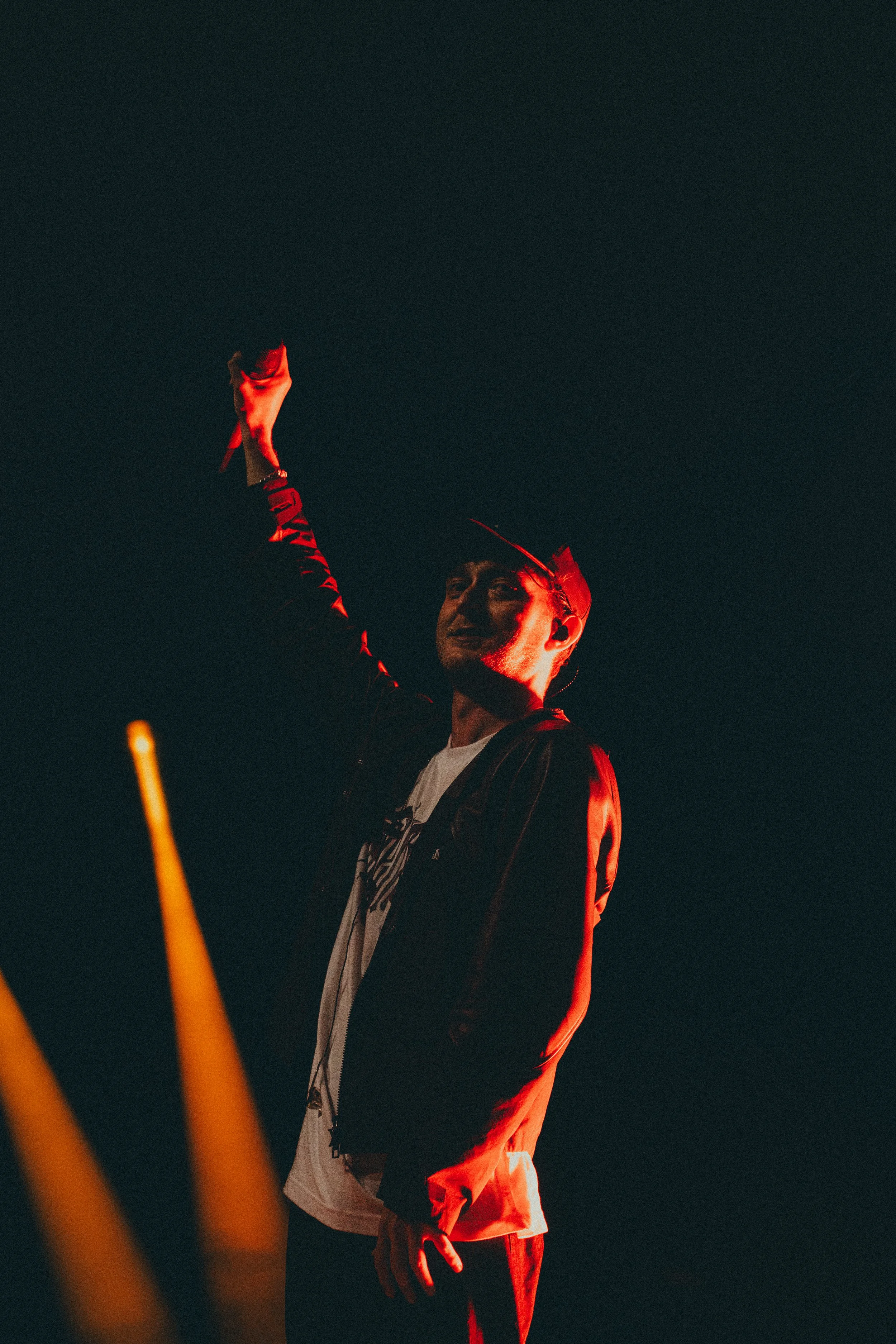 Une personne debout dans l'obscurité, avec un éclairage rouge, qui l'éclaire partiellement. La personne porte une casquette et un veste en cuir, et elle semble faire un geste avec la main levée.