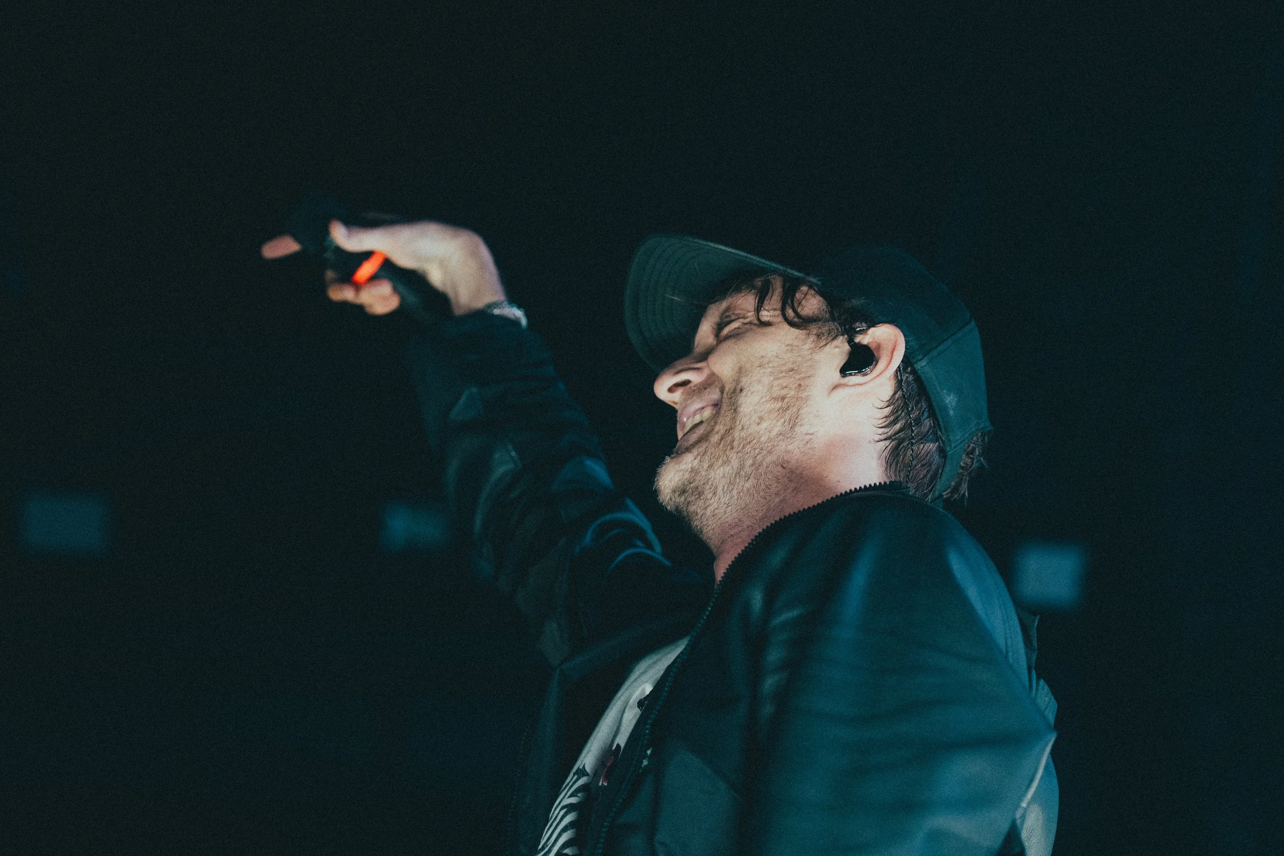 Un homme souriant, portant une casquette et une veste en cuir, semble être à un concert ou un événement, tenant un objet dans sa main.