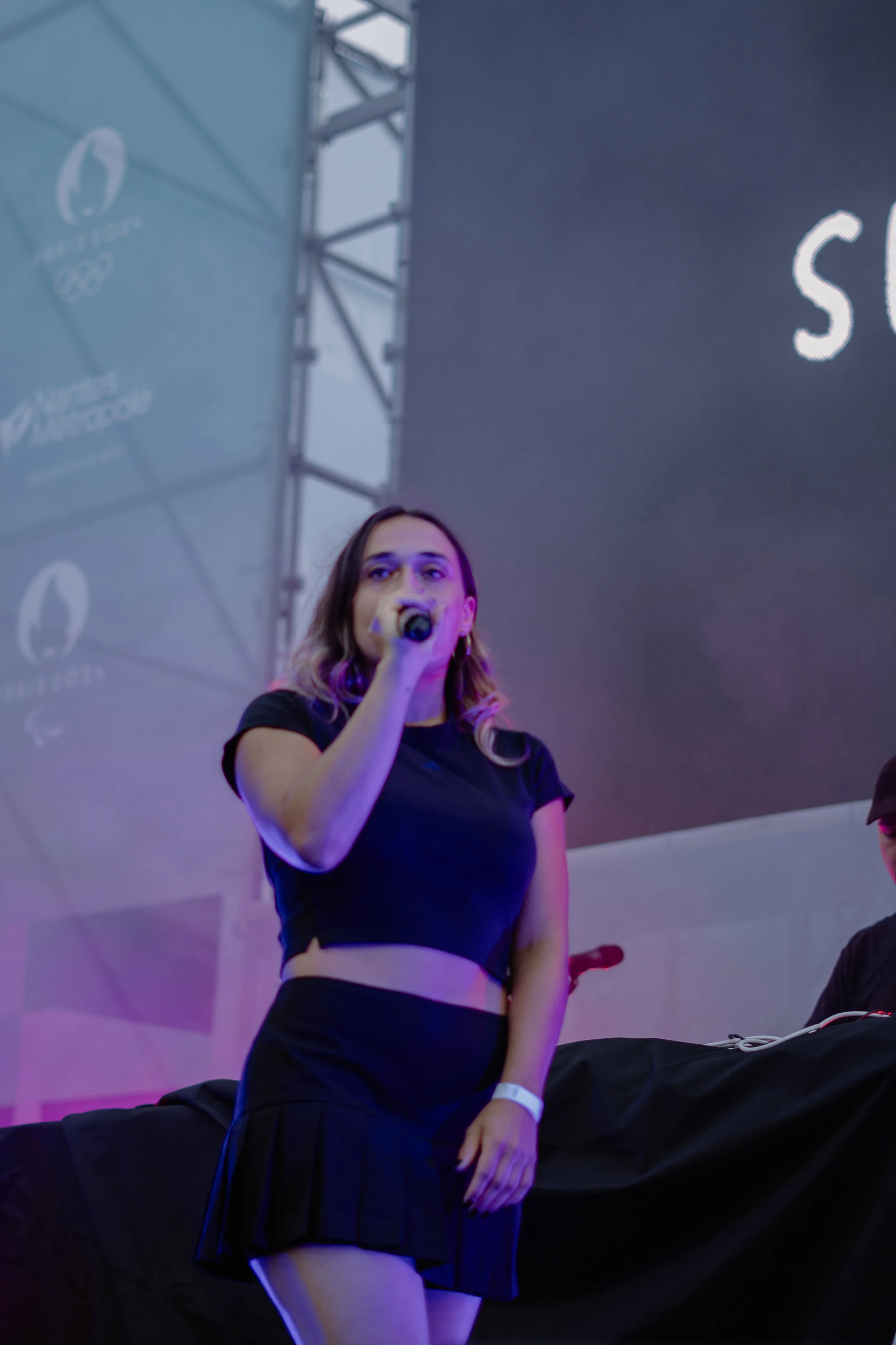 Une jeune femme tient un micro lors d'un événement, avec un écran géant derrière elle et un acteur en arrière-plan.