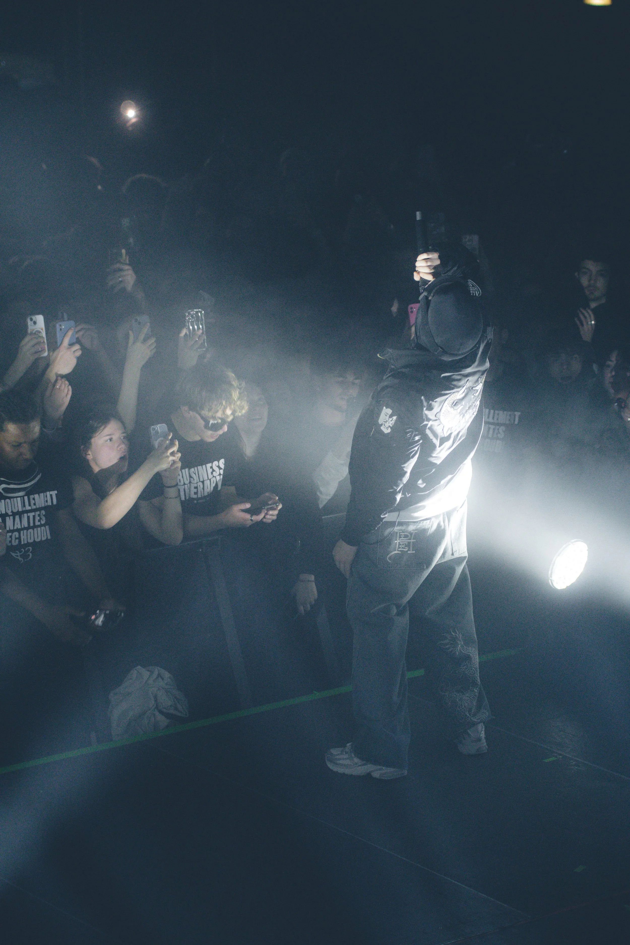 Un artiste de hip-hop sur scène dans une lumière dramatique, entouré de fans prenant des photos et vidéos avec leur téléphone dans une ambiance sombre.