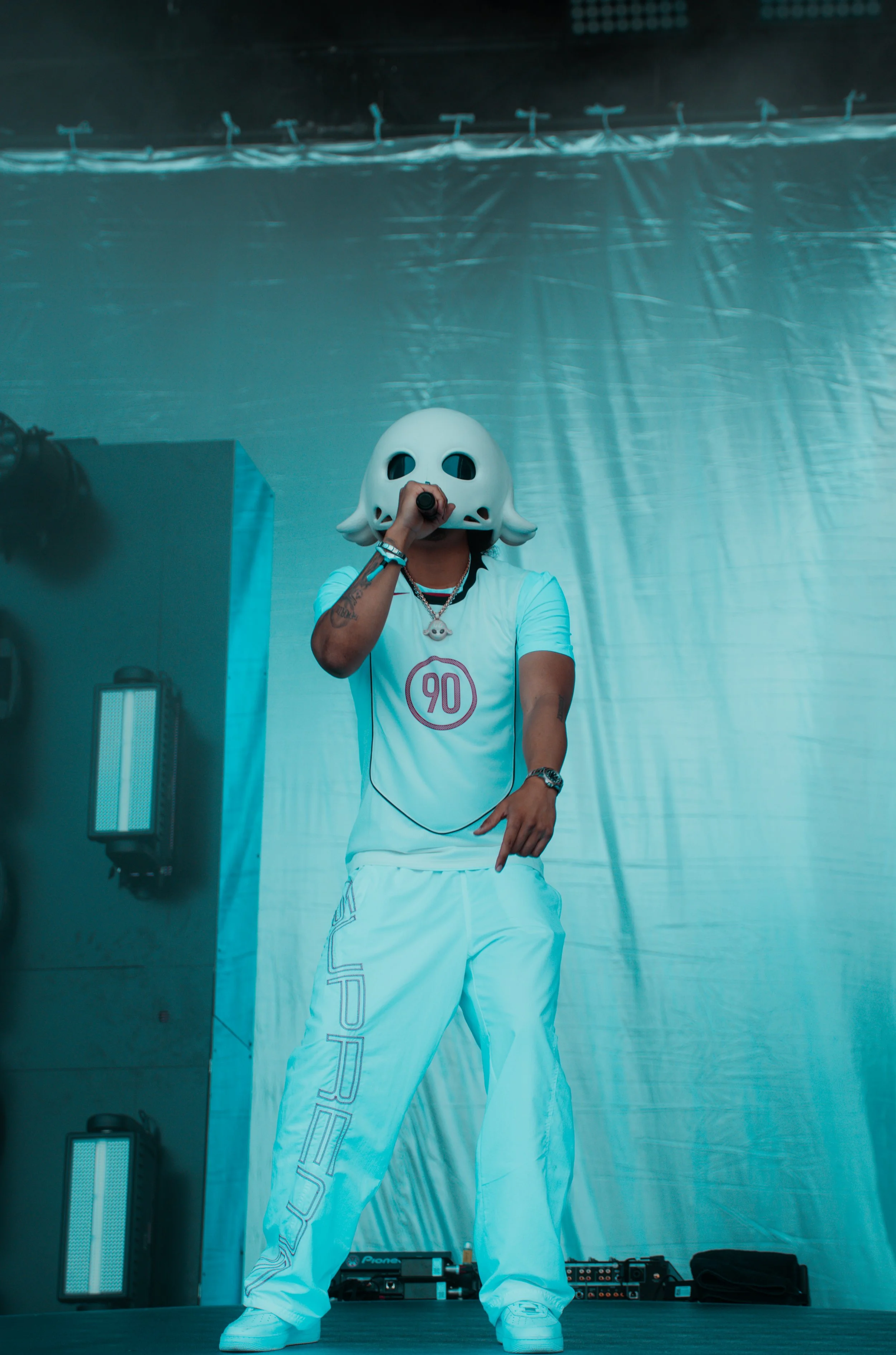 Un artiste portant un masque blanc en forme de visage de cartoon, habillé en tenue blanche, en train de performer sur scène avec un microphone.